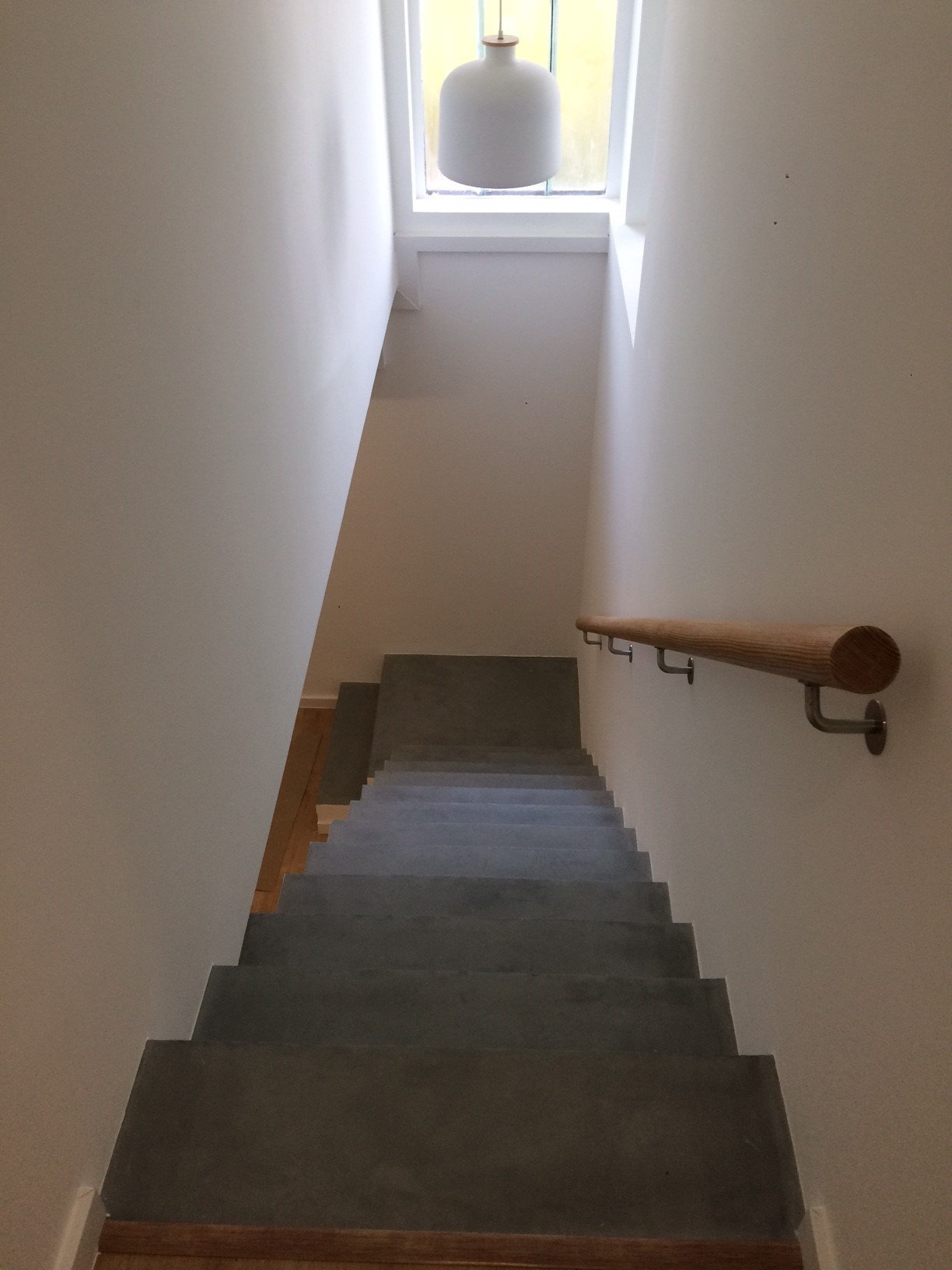Escada de uso interno vista de cima, com degraus de pedra, parede branca ao lados, uma janela ao fundo e uma luminária branca pendurada no teto.