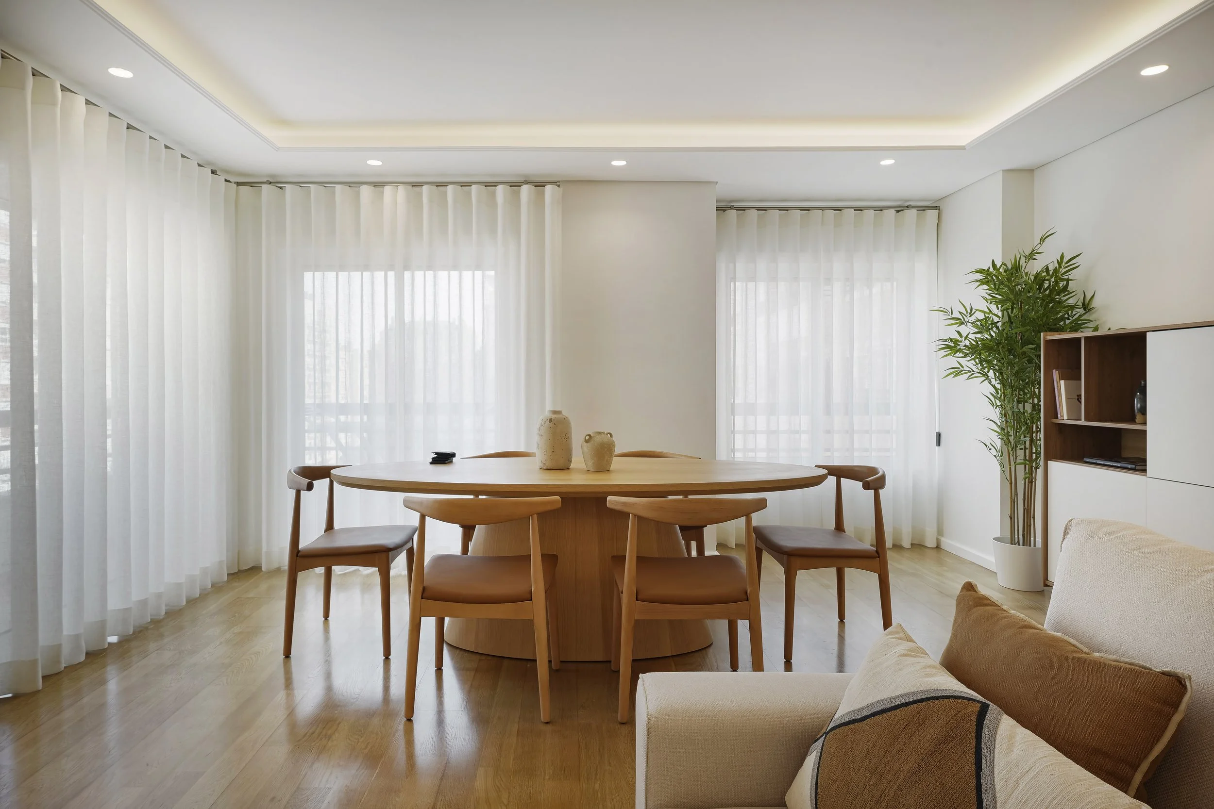 Sala de jantar minimalista com mesa de madeira oval, seis cadeiras de madeira com assentos em pele sintética, cortinas brancas transparentes.