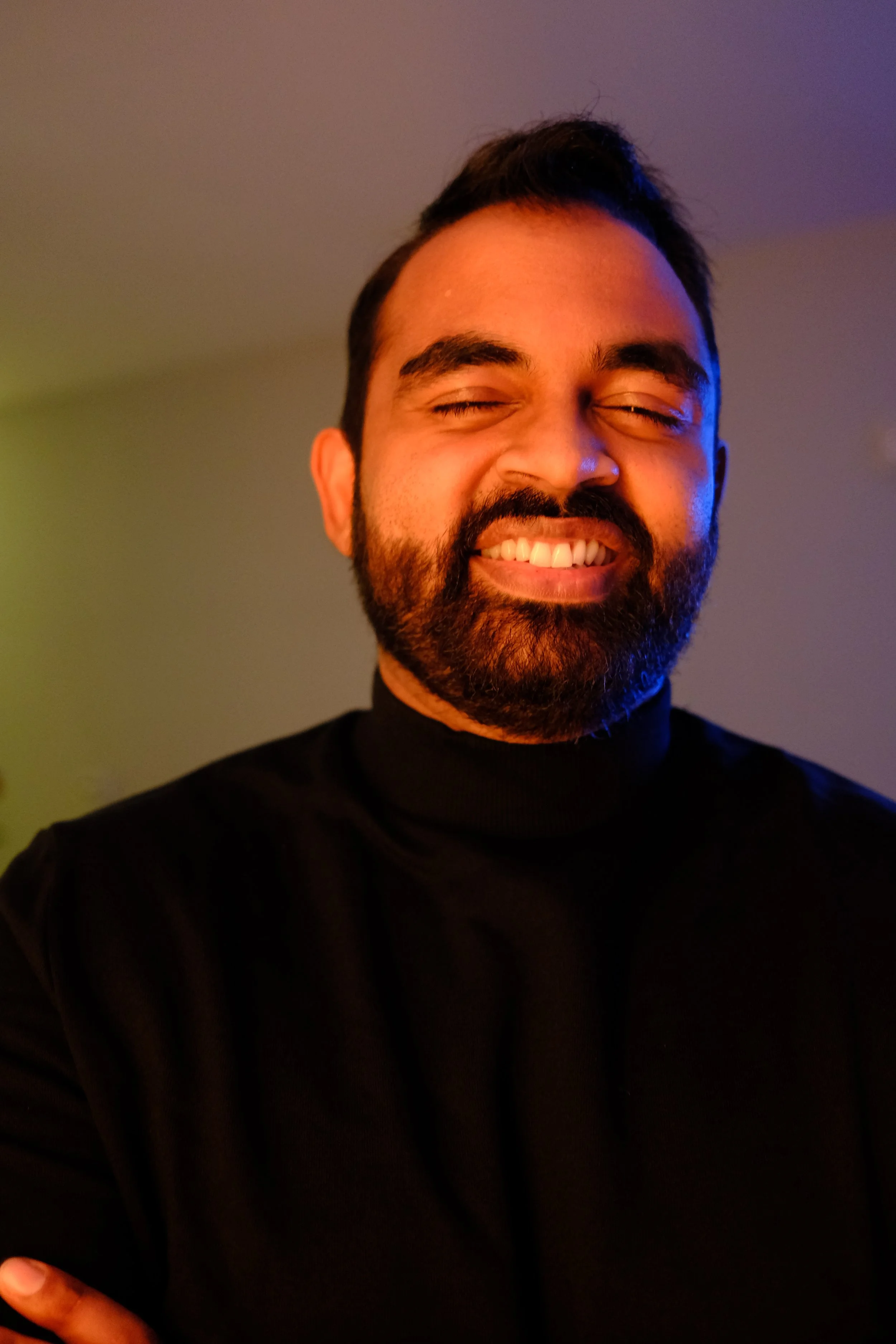 A man with a beard smiling with his eyes closed, wearing a black turtleneck, against a dim colorful background.