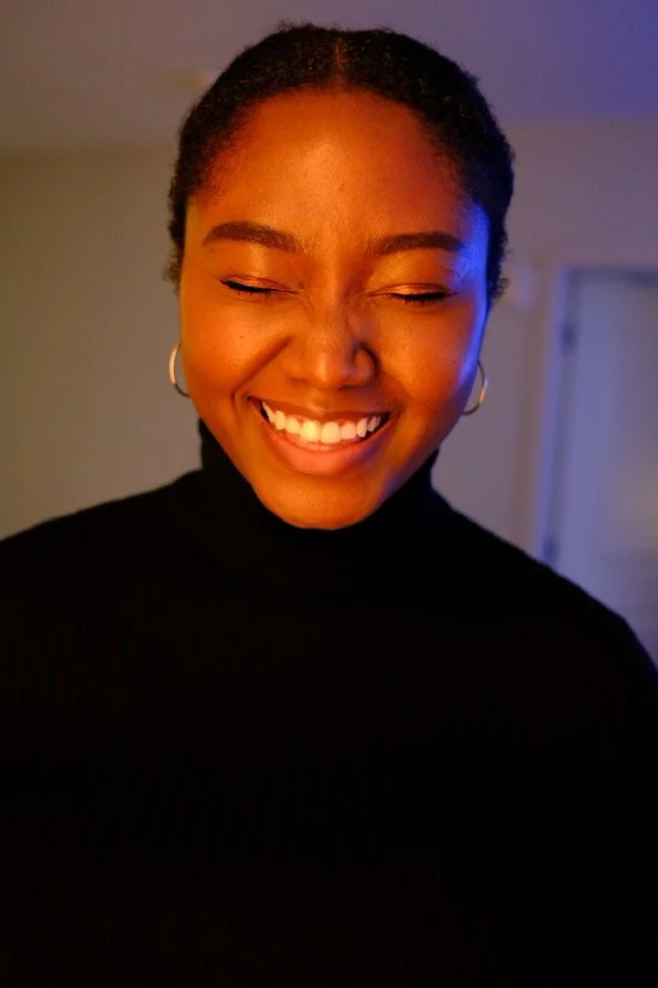 A woman with dark hair pulled back, smiling with her eyes closed in a black turtleneck, standing indoors with a neutral background.