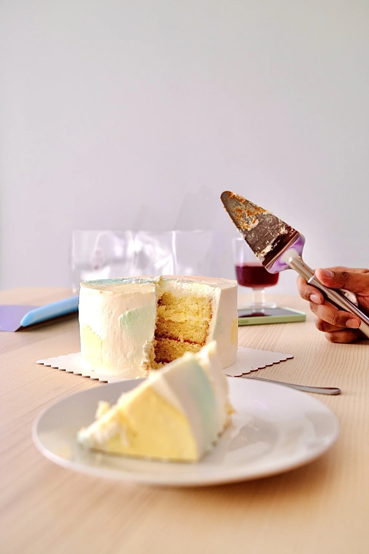 A sliced cake with white icing on a table, with a hand holding a cake server and a glass of red wine in the background.