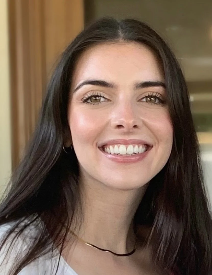 Close-up of a woman with long, dark hair, smiling, showing teeth, wearing a white top, a thin black choker, and small hoop earrings.
