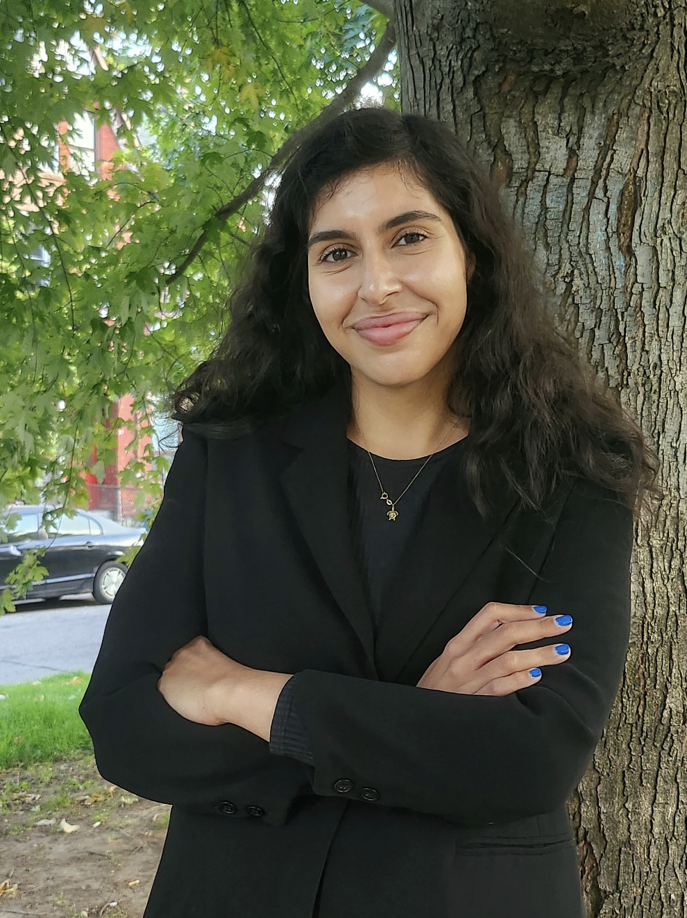 A woman with curly dark hair and a black blazer standing outdoors by a tree with green leaves, crossing her arms and smiling.