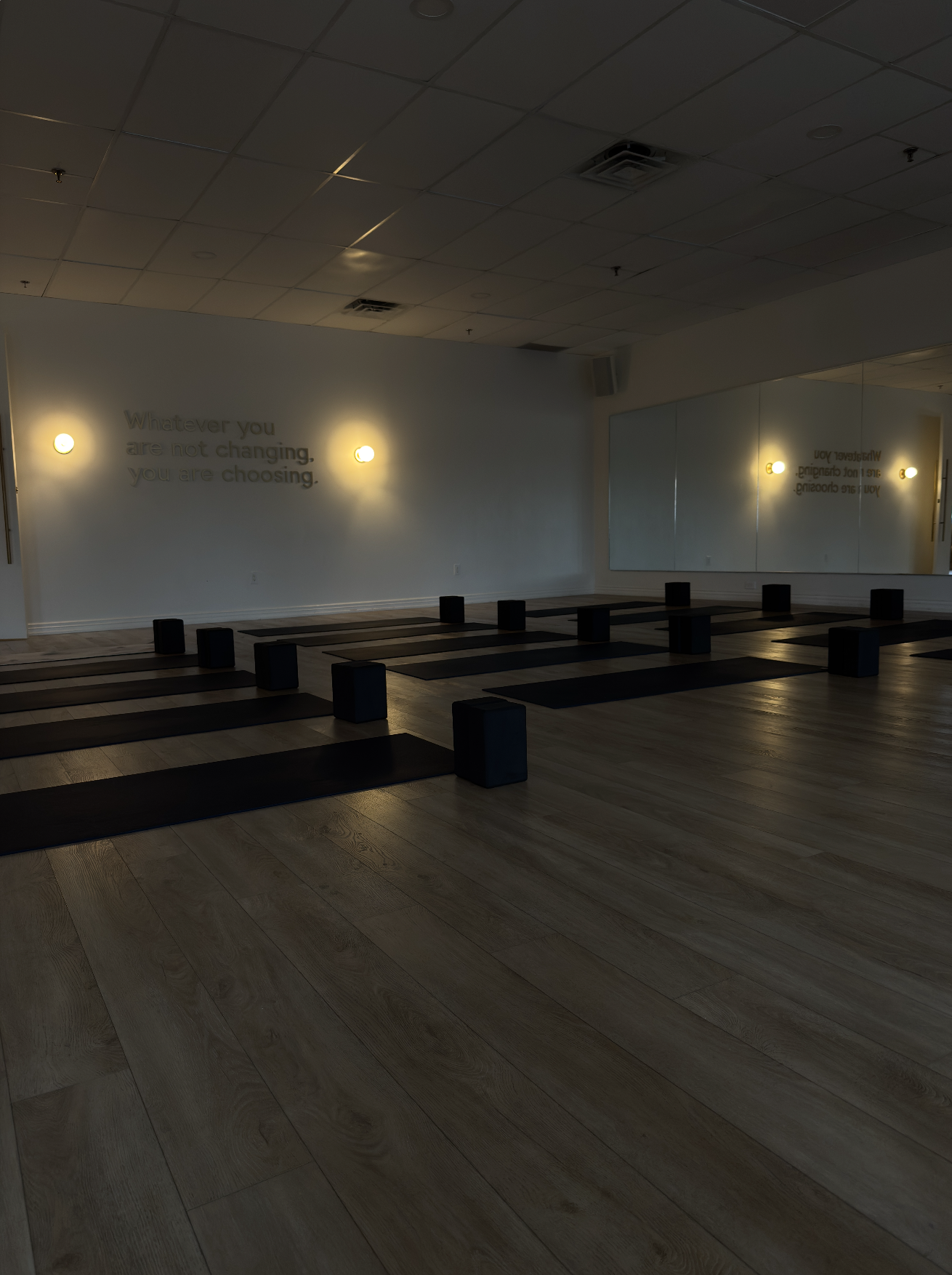 Yoga studio with black mats and bolsters arranged on wooden floor, mirrored wall, dim lighting, and wall-mounted lights