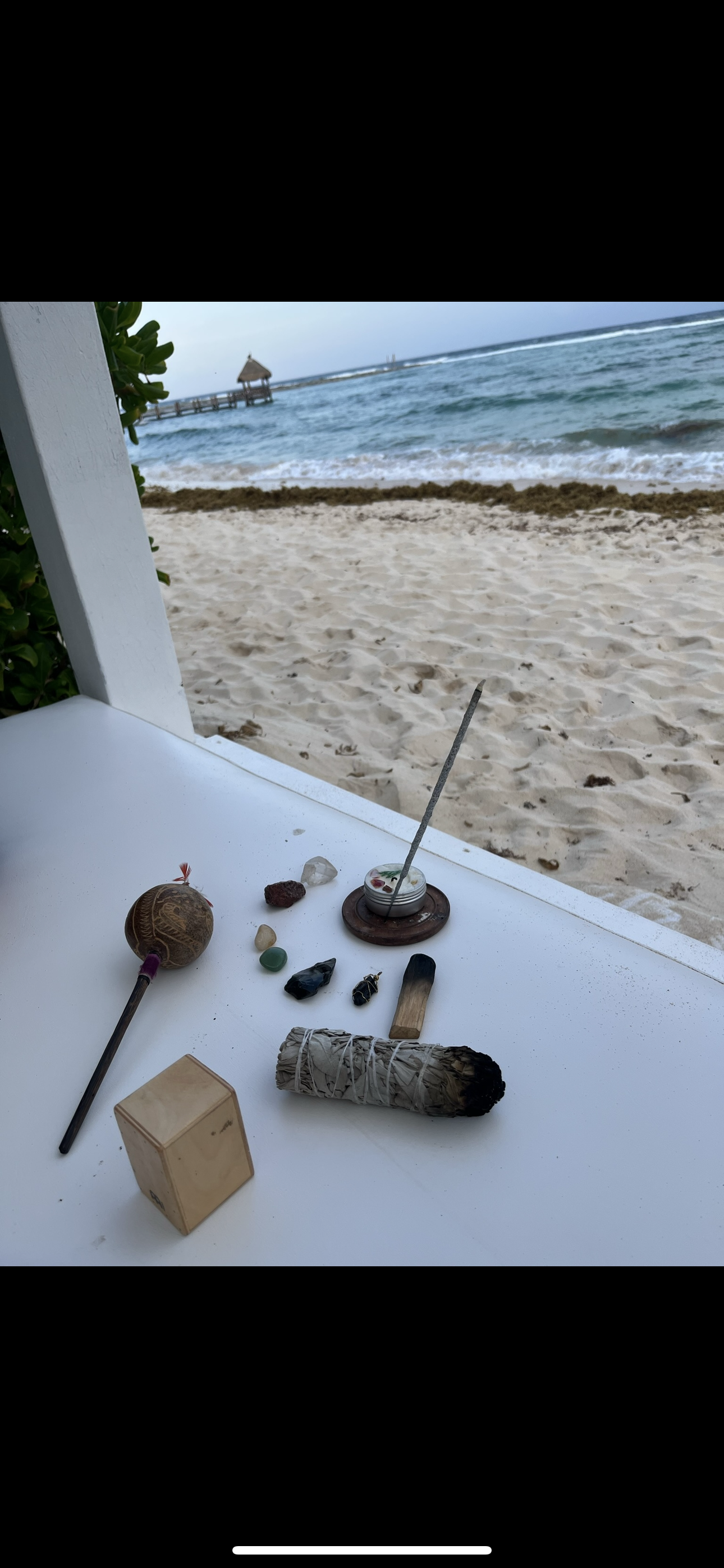 Collection of spiritual and witchcraft items on a white table by the beach, including a bundle of sage, crystals, a bowl with a wand, a wooden box, and a black stone, with ocean and a pier in the background.