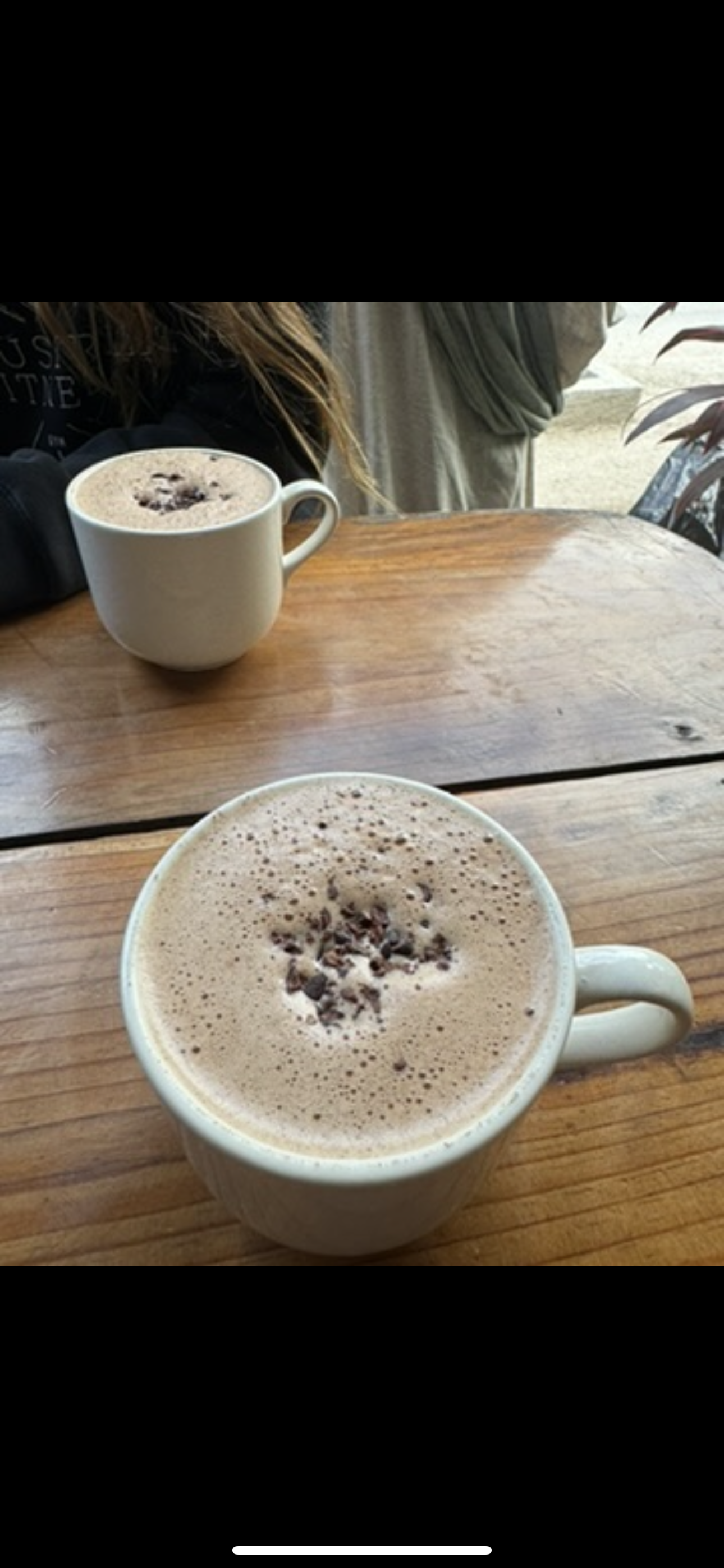 Two cups of hot chocolate topped with chocolate shavings sit on a wooden table in a cozy setting.