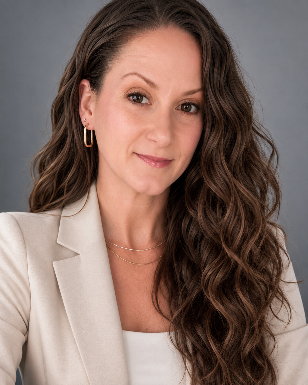 Headshot of a woman with long wavy brown hair, wearing a beige blazer, gold hoop earrings, and layered necklaces, against a gray background.