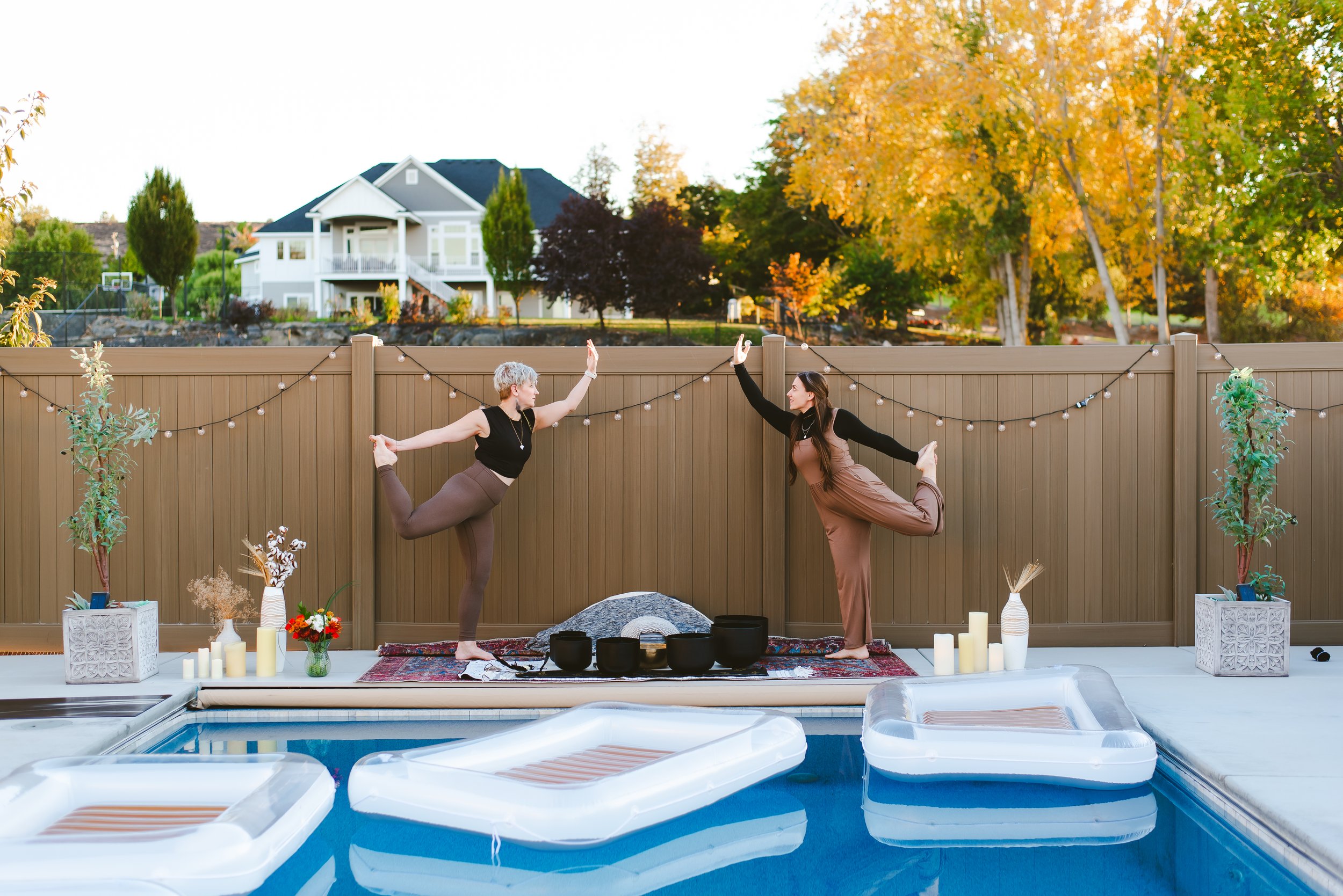 Two women standing in front of a fence holding yoga poses, with a pool in the foreground. Yoga, sound bath, and breathwork session.