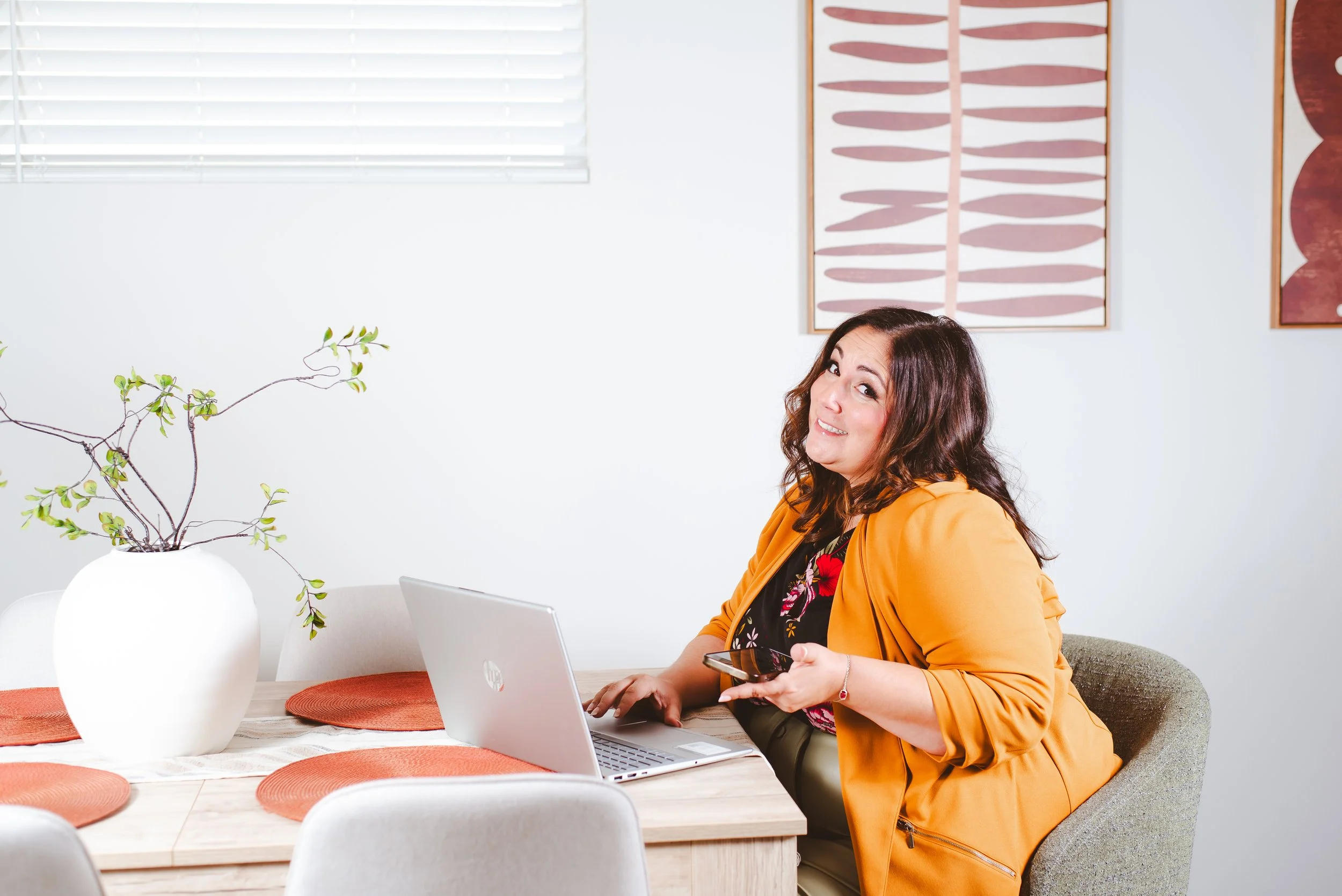 Woman sitting at a kitchen table with 1 hand on a laptop keyboard and the other holding her phone - realtor life - shrugging her shoulders and smiling at the camera.