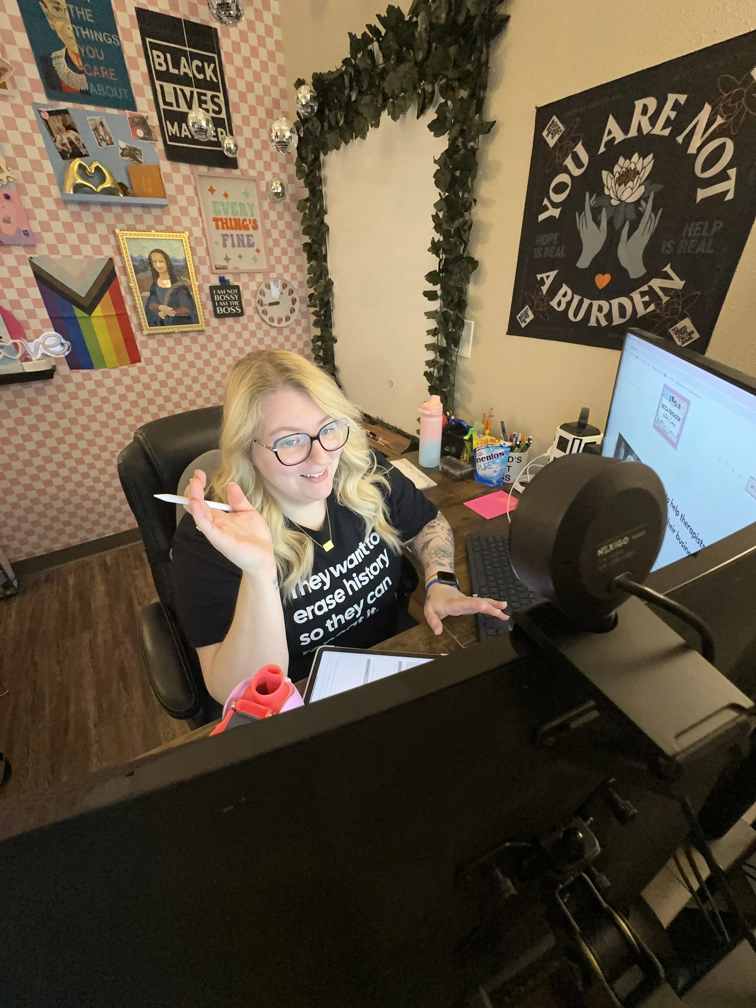 A woman with blonde hair, glasses, and a black T-shirt sitting at a desk in front of a computer, smiling and holding a pen. The room's wall behind her has various colorful posters and artwork, including a rainbow flag and a 'Black Lives Matter' sign. There is a large black poster with white text that reads 'You are not a burden,' surrounded by decorative elements. The desk has a tablet, a pink water bottle, snacks, and stationery.