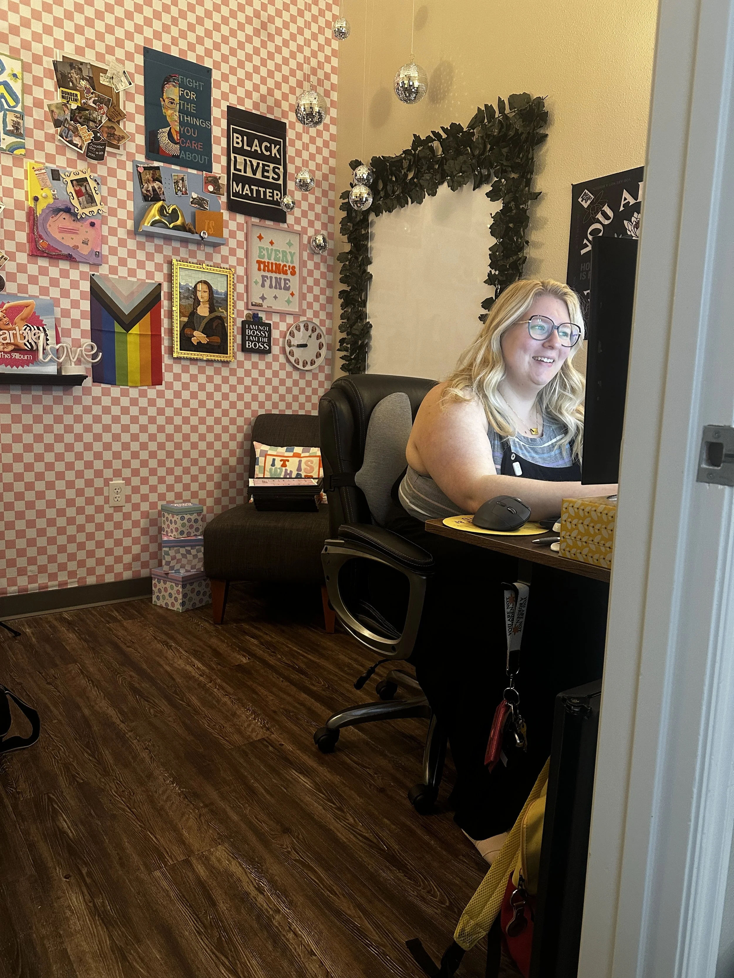 A woman with blond hair and glasses working at a desk with a computer in an office decorated with various posters, signs, and wall art, including a Black Lives Matter sign, Mona Lisa, and rainbow pride flag. The Therapy Office, Springdale, AR
