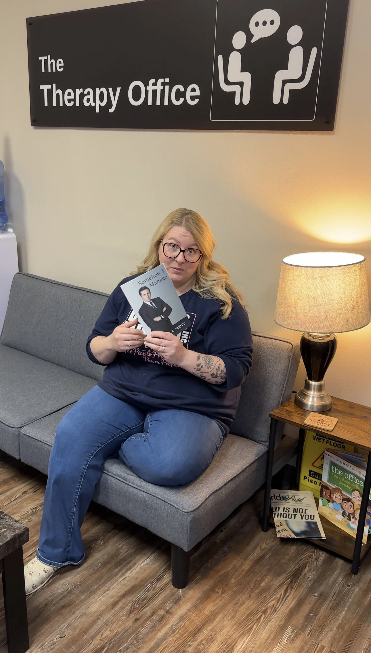A woman sitting on a gray couch in a therapy office, holding a book, with a sign reading 'The Therapy Office' and an illustration of two people talking on the wall behind her, and a small table with a lamp and magazine nearby. The Therapy Office, Spr