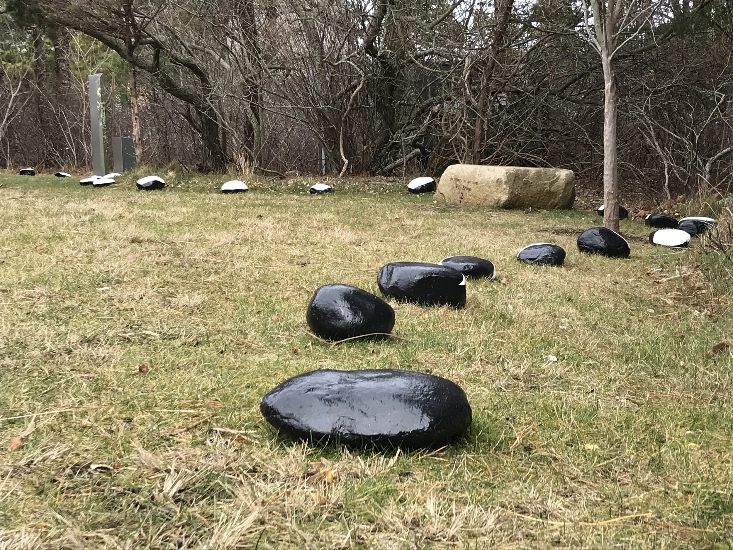 A pathway lined with black and white painted rocks on grass, with a stone and trees in the background.
