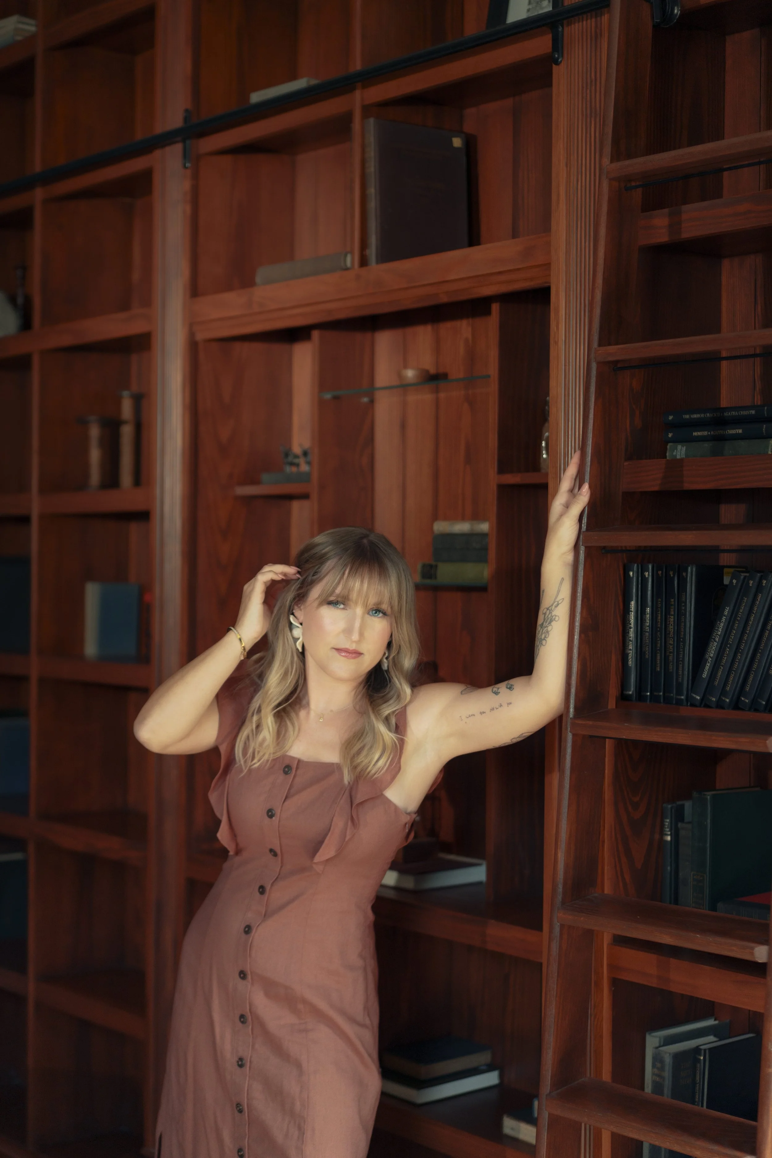 A blonde business owner with long blonde hair leans against a beautiful mahogany library backdrop, tucking her hair behind her ear. She looks directly at the camera, inviting the viewer to join the experience.