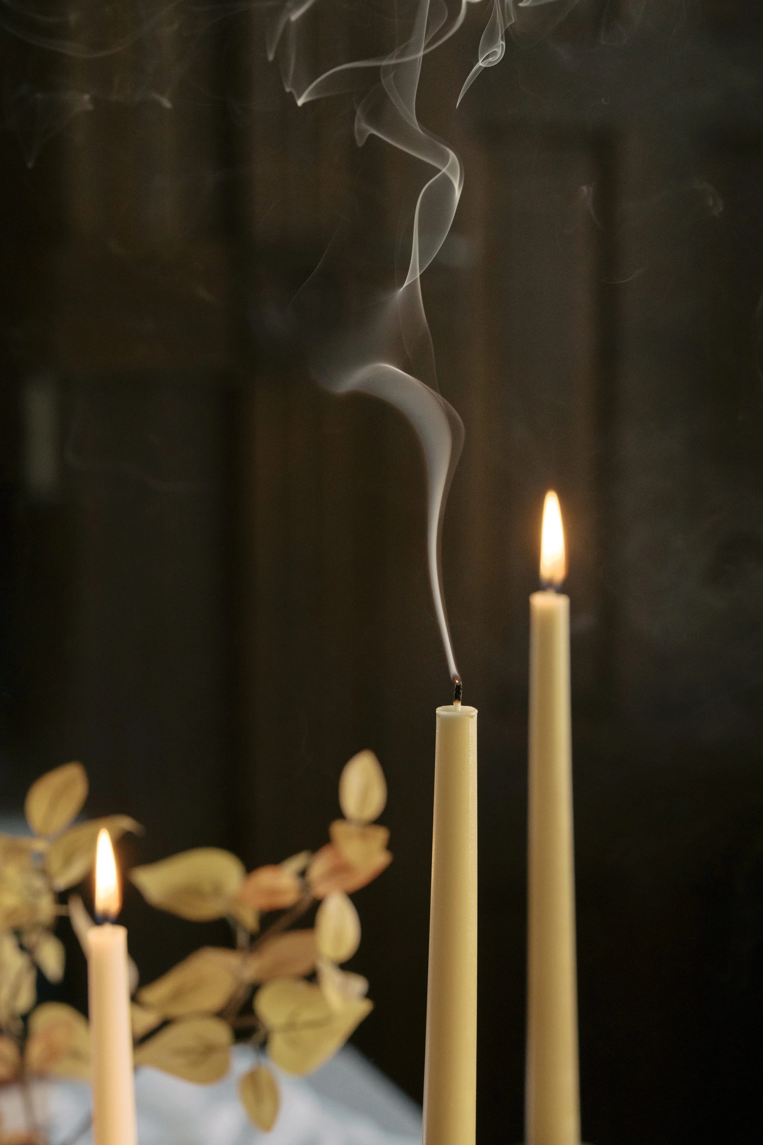 Against a dark backdrop, three candles rise from the bottom of the image. Two are lit while the middle candle extends a smoke trail, just extinguished.