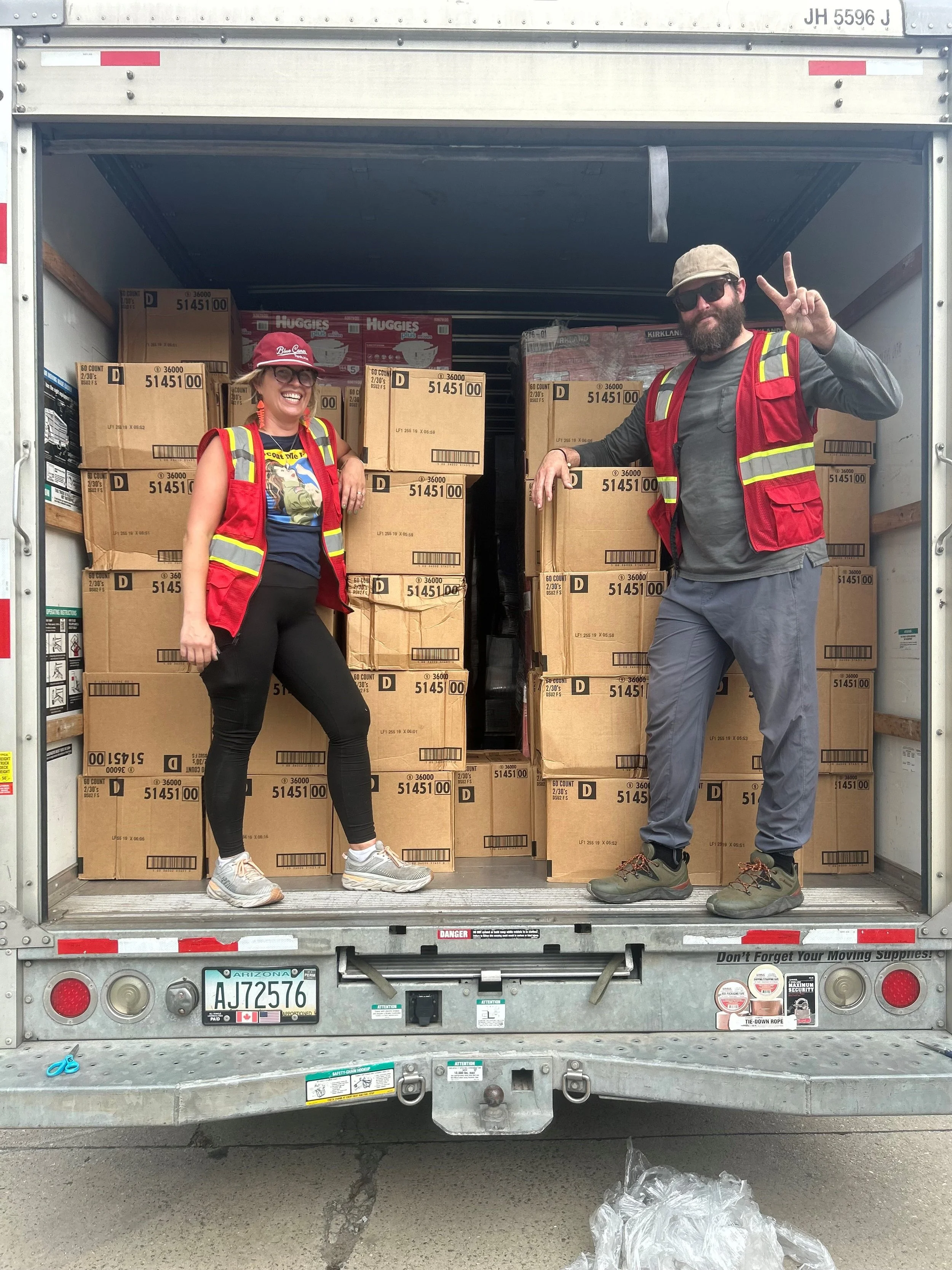 Two people wearing red safety vests standing in the back of a truck filled with boxes of diapers, smiling and posing for the photo.