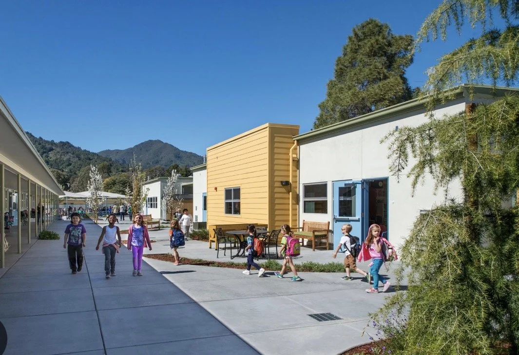 Elementary school campus with children walking between colorful buildings and forested mountains under a clear blue sky