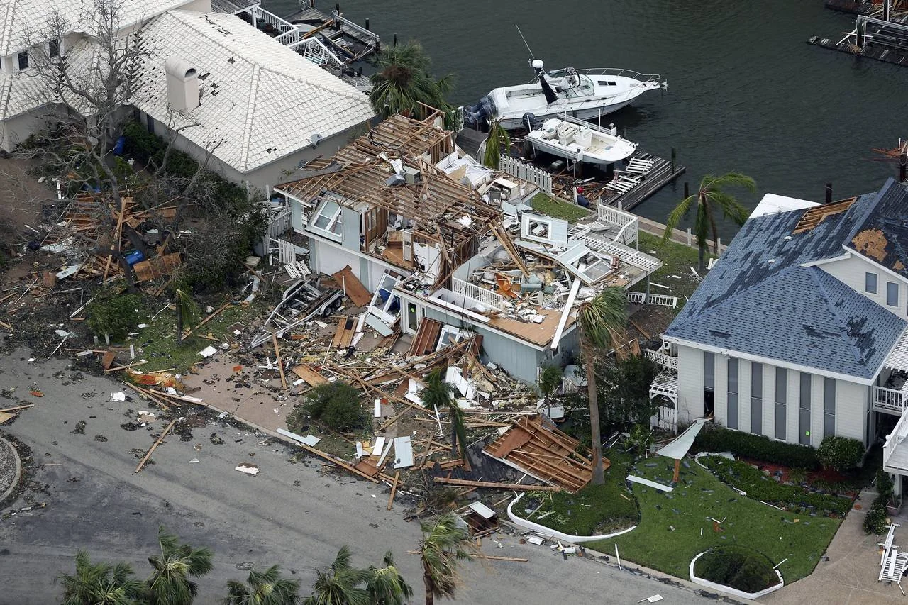A residential neighborhood with significant damage from a storm or natural disaster. One house's roof and interior are heavily wrecked, with debris scattered on the ground. There is a neighboring house with a intact roof, and boats are docked along the water nearby.