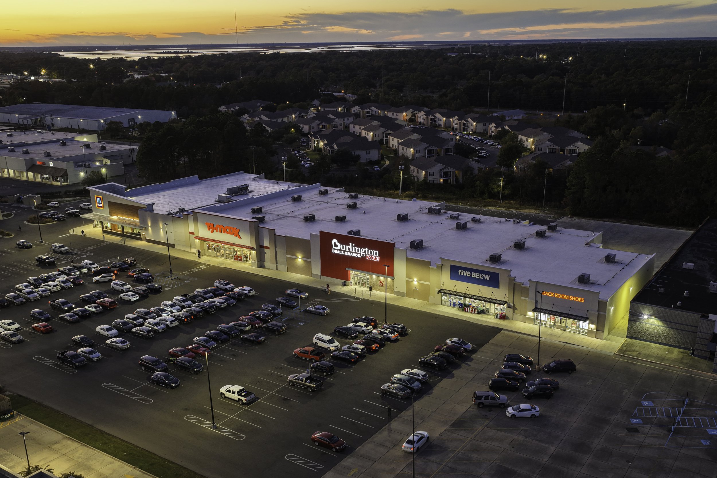 An aerial view of a shopping plaza at dusk featuring a parking lot filled with cars, a T.J. Maxx store, Burlington, Five Below, and Black Room Shoes stores, with a residential area and water body in the background.