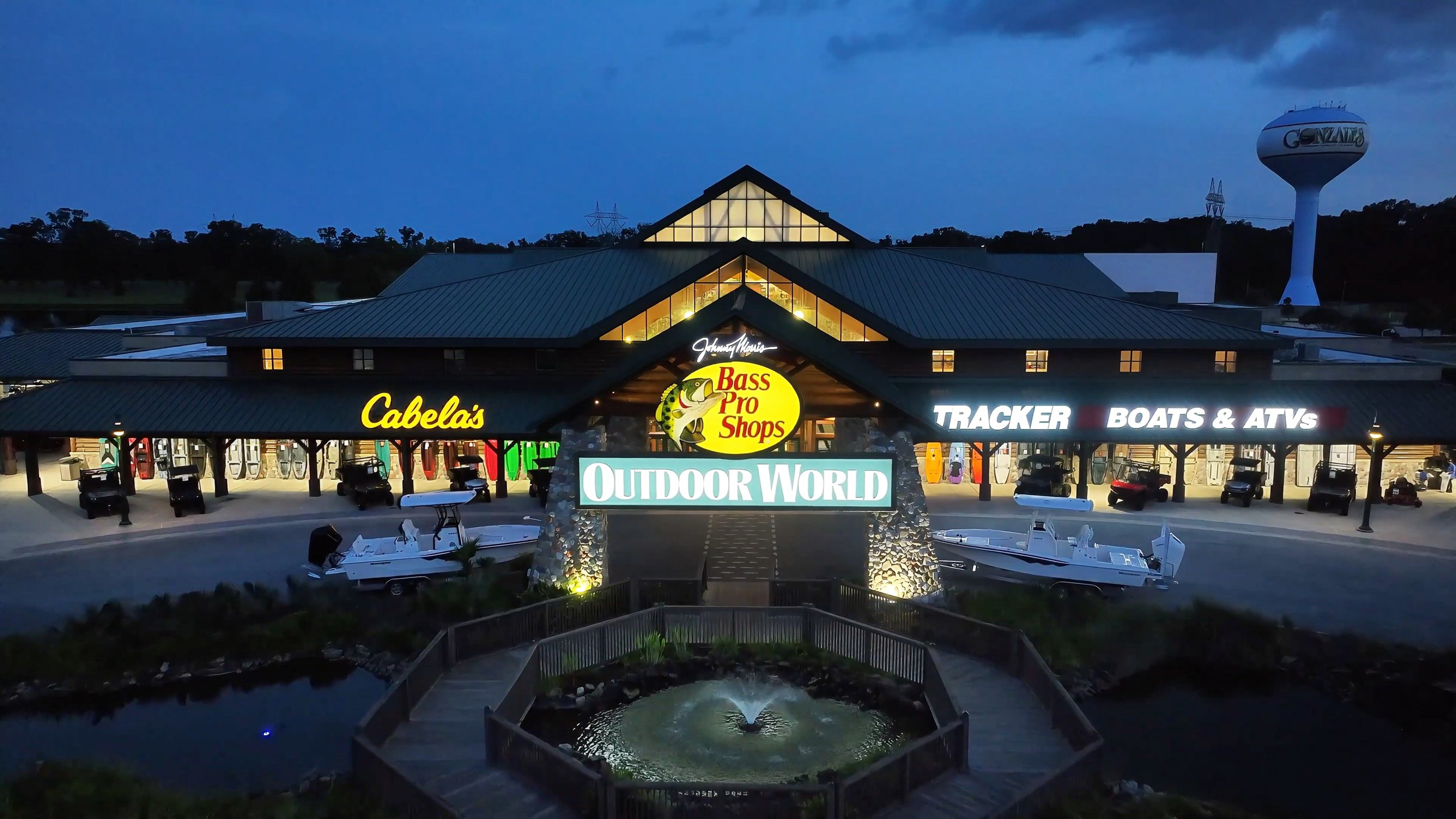 Nighttime view of Outdoor World store with illuminated signs for Cabela's, Bass Pro Shops, Tracker boats, and ATV rentals, featuring boat displays outside and a water fountain in the foreground.