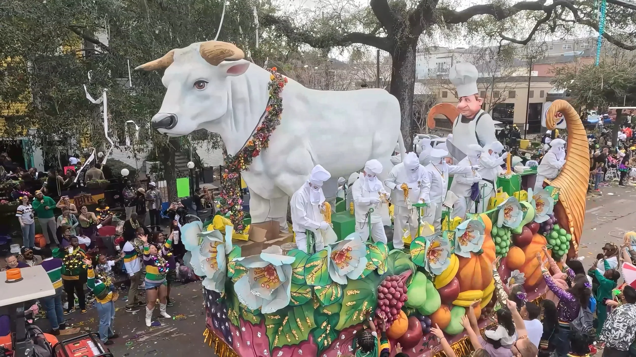 A large parade float featuring a giant white bull with a flowered collar, a chef figure, and a snake coiled around the float, surrounded by people in festive attire and a large crowd.
