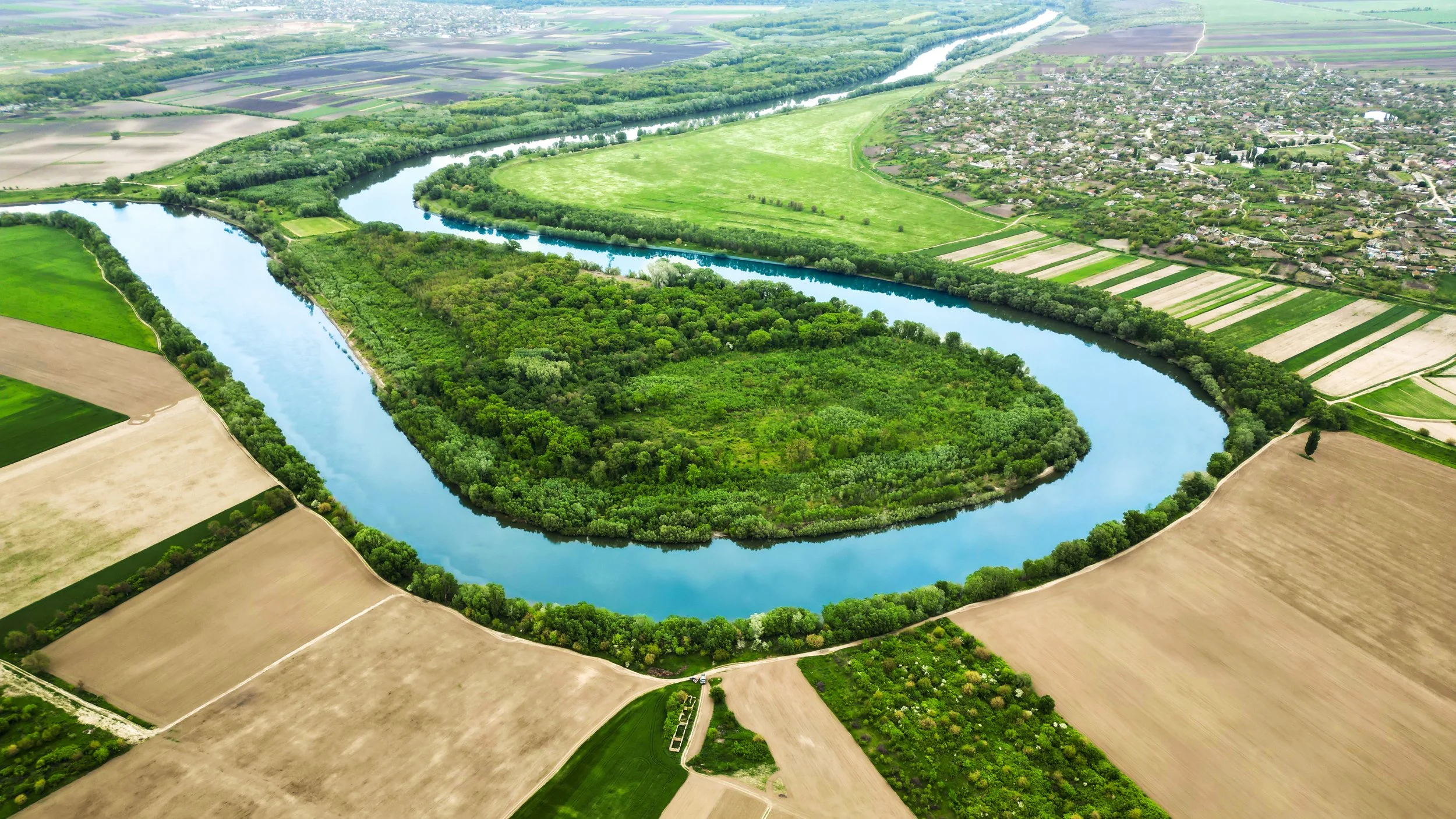 An aerial view of a winding river with surrounding green trees and farmland, adjacent to a residential area.