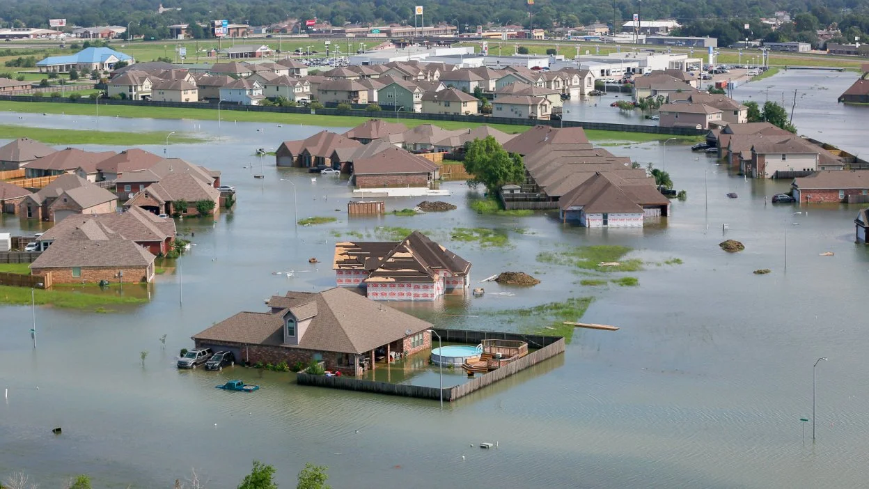 A neighborhood flooded with water covering yards, driveways, and streets, with houses surrounded or partially submerged, and debris floating on the water.
