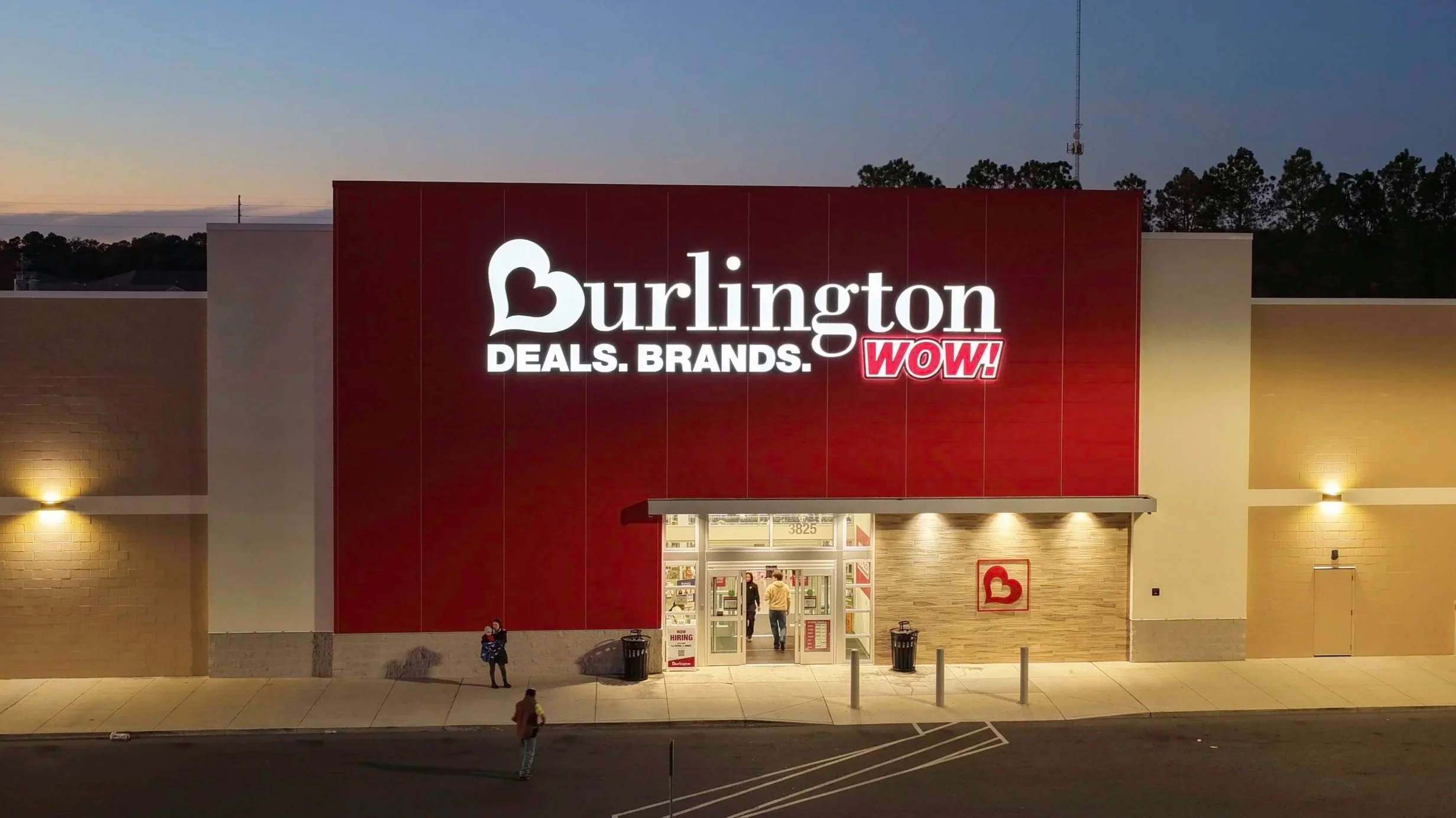 Exterior of Burlington retail store during evening with illuminated sign reading 'Burlington DEALS. BRANDS. WOW!' and people entering and exiting the store.