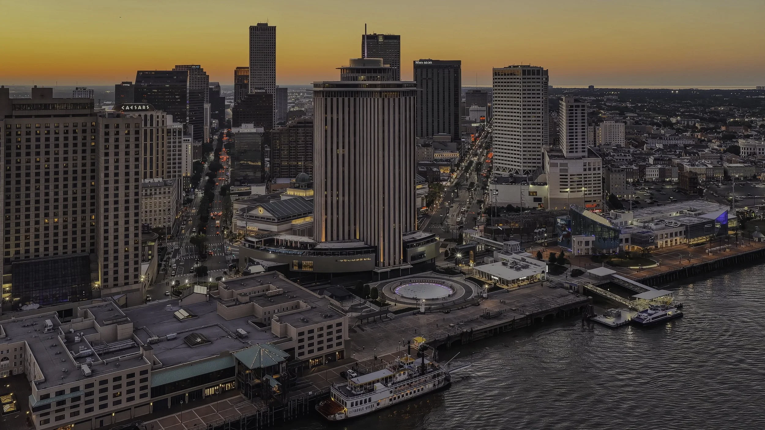Aerial view of a city skyline at sunset near a harbor, with tall buildings, a pier, and boats docked in the water.