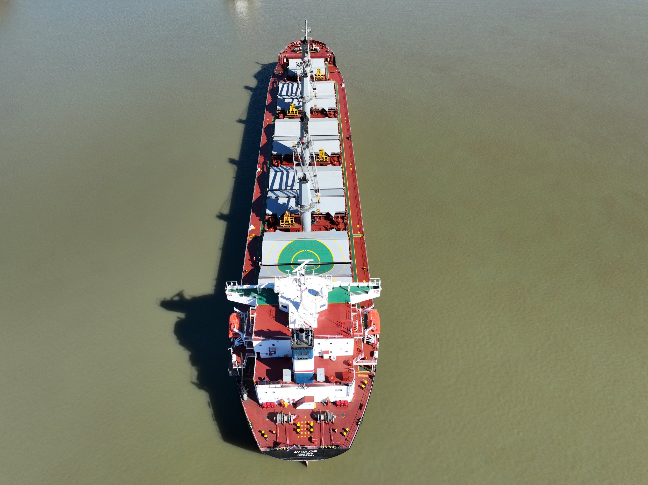 An aerial view of a large red and white military ship with a helicopter pad, sailing on calm water.
