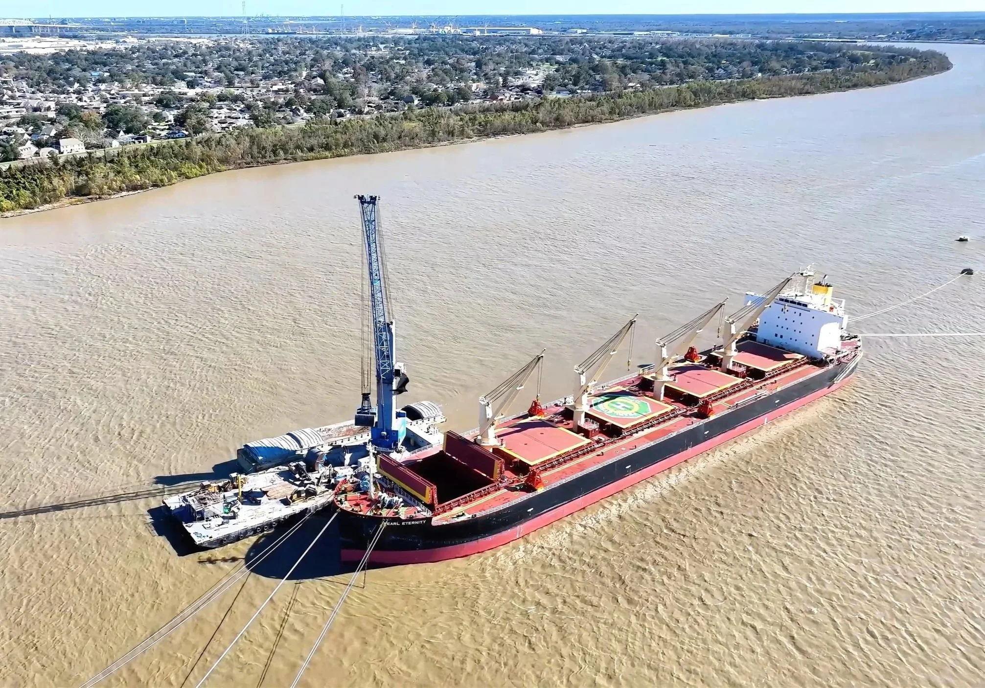 Large cargo ship named Pearl Eternity with four cranes on deck, anchored in a muddy river near a city shoreline.