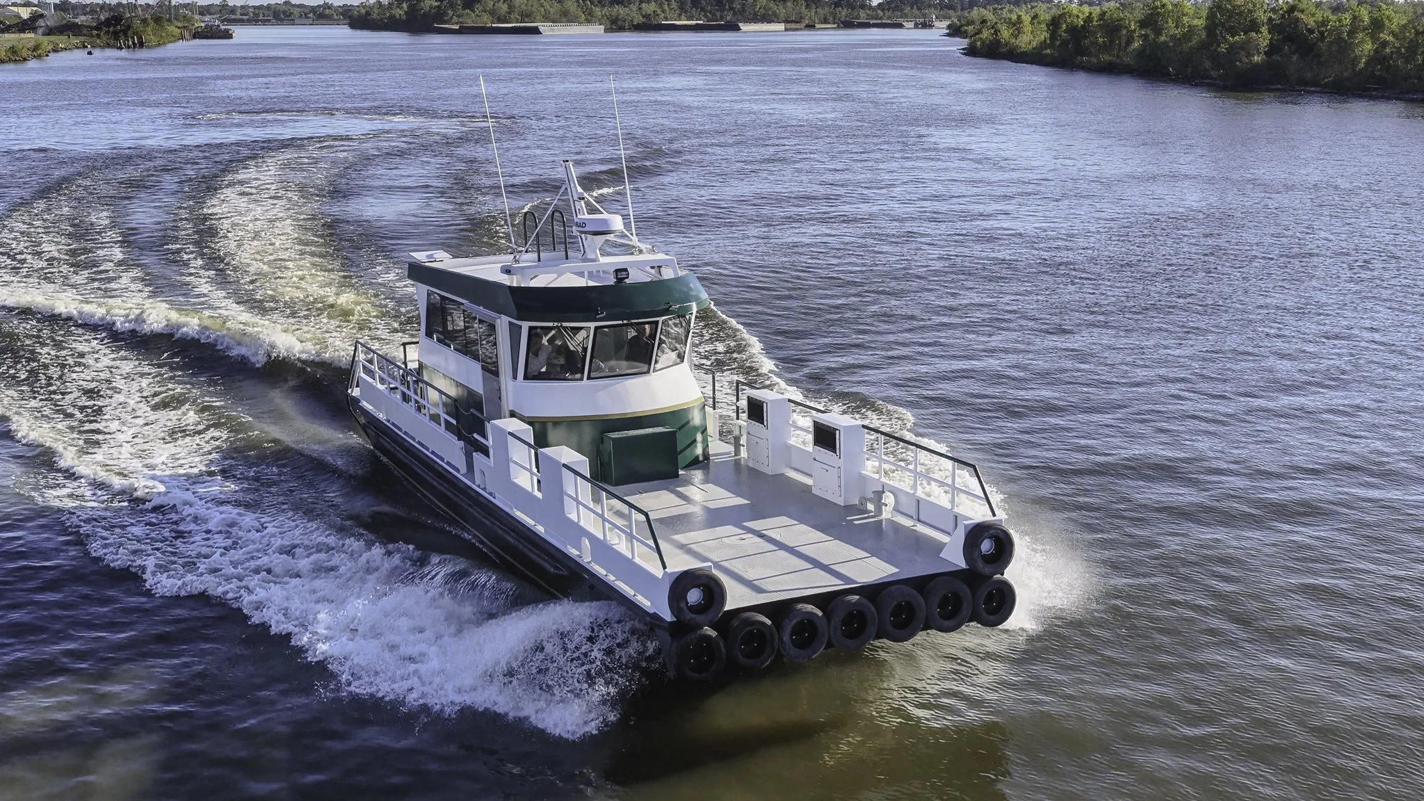 A small motorboat moving through a body of water, creating waves behind it. The boat has a covered cabin area and is outfitted with fenders around its sides. Trees line the shoreline in the background.