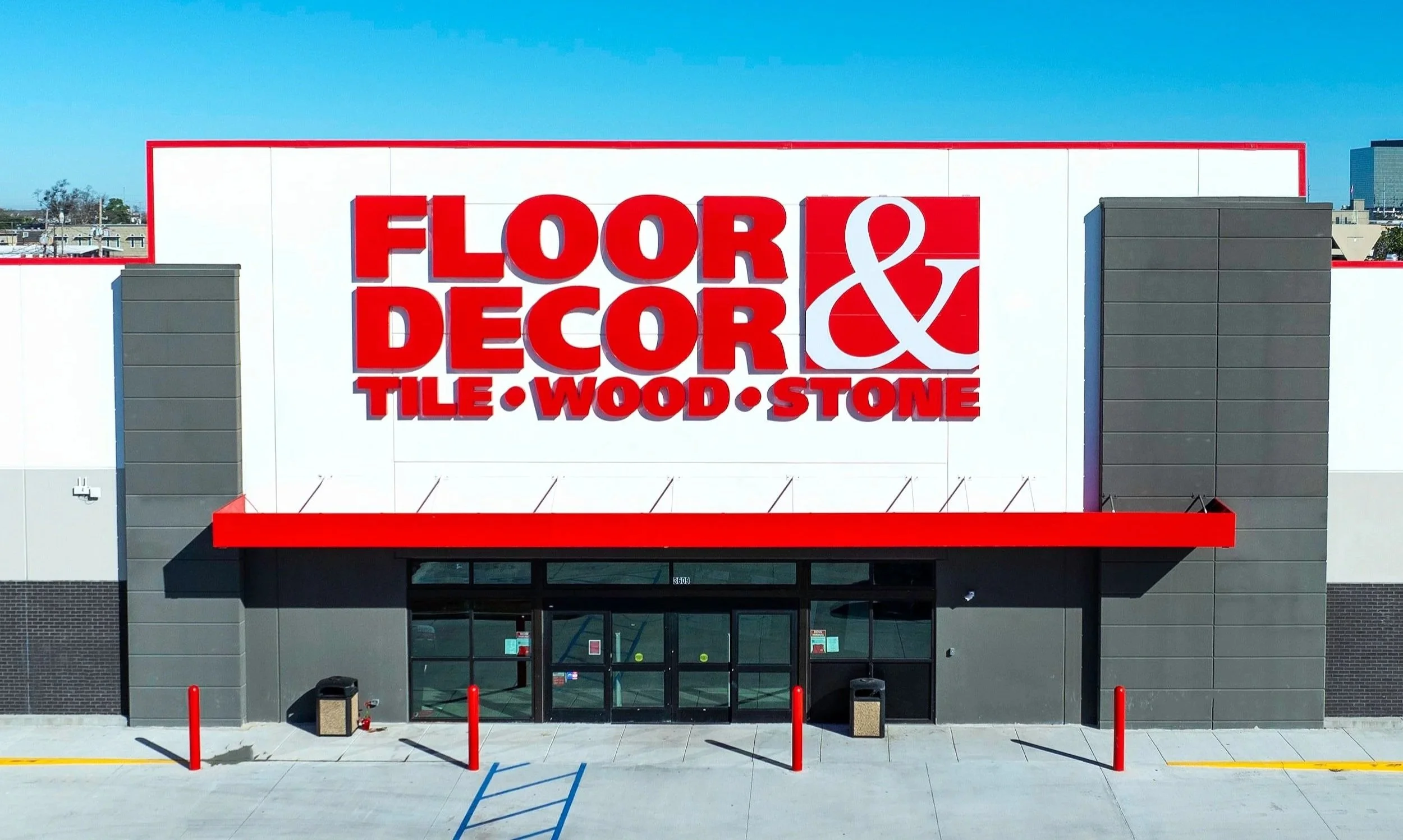Exterior of a store with large sign reading "FLOOR & DECOR TILE, WOOD, STONE" in red letters on a white background, gray walls, and a glass entrance door, with a clear blue sky above.