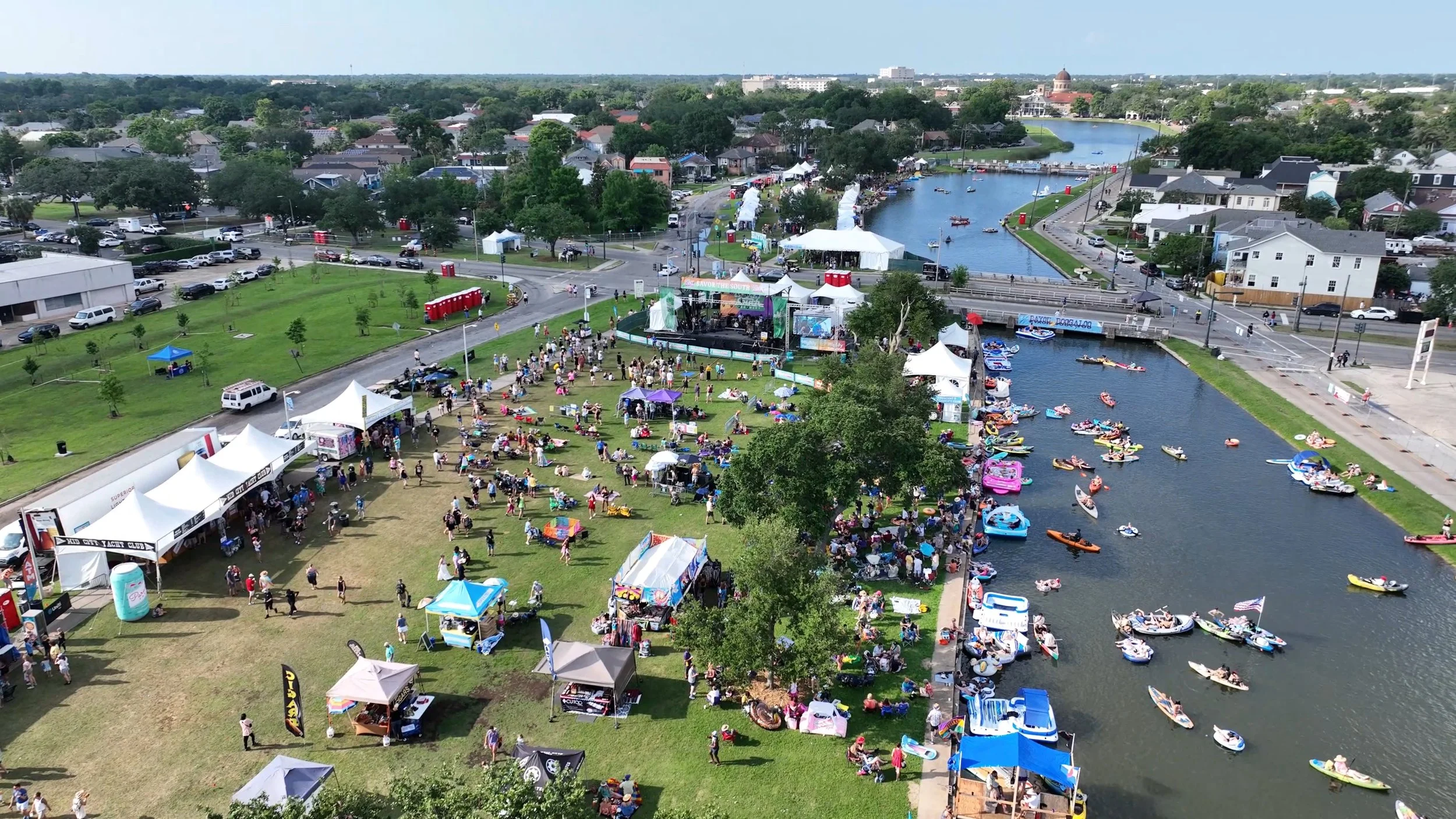 Aerial view of a lively outdoor festival by the water with tents, a stage, and many people. Boats are on the river, and there are green spaces, trees, and a cityscape in the background.