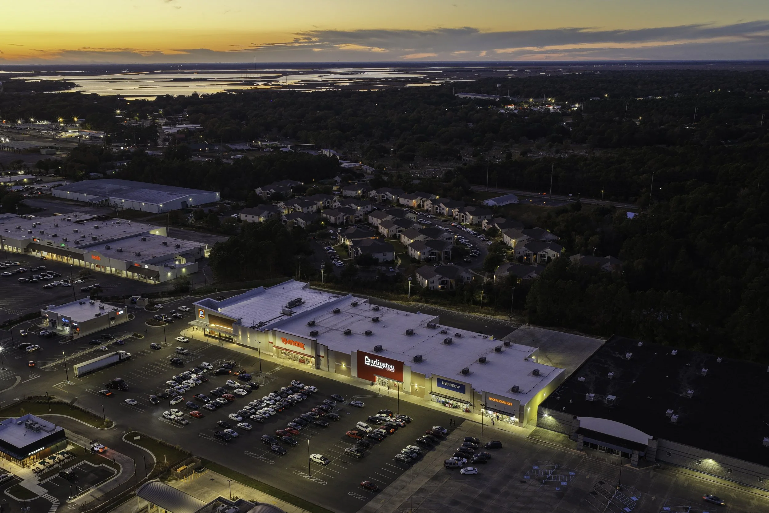 Aerial view of a shopping center parking lot at dusk, with stores including Burlington, T.J. Maxx, and Five Below, and a residential neighborhood in the background.