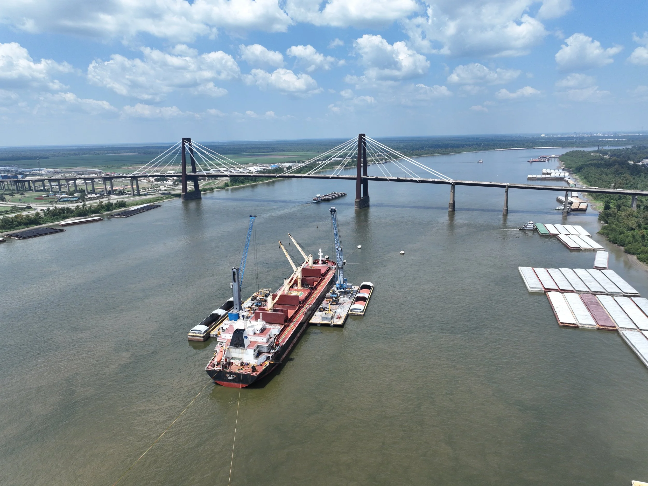Aerial view of a river with a large cargo ship docked, crane equipment on the ship, and a bridge in the background under partly cloudy sky.