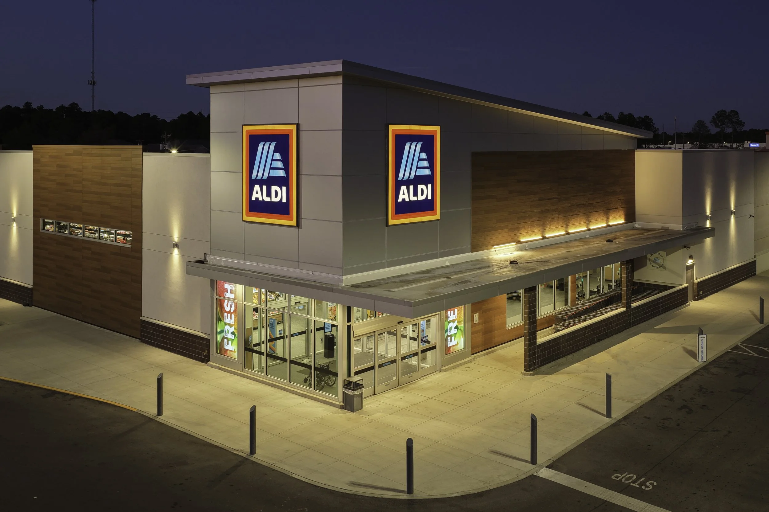 An Aldi grocery store at night with illuminated signs and entrance, surrounded by a paved parking area with a stop sign.