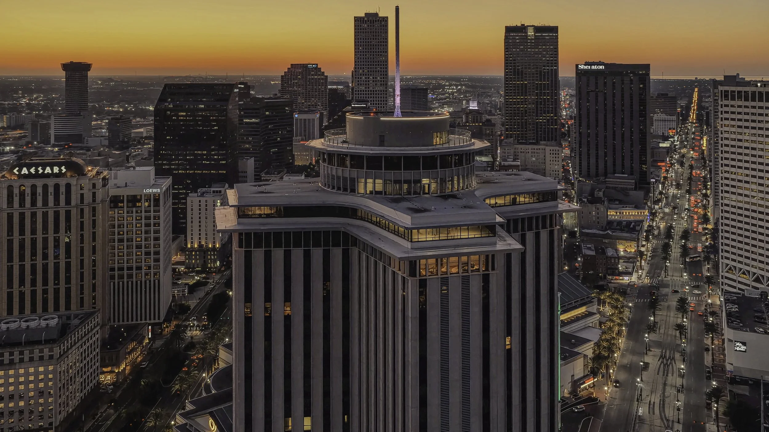 Aerial view of a city skyline at sunset with tall buildings, including Caesars, Loews, and Sheraton hotels, and streets filled with cars and illuminated by streetlights.