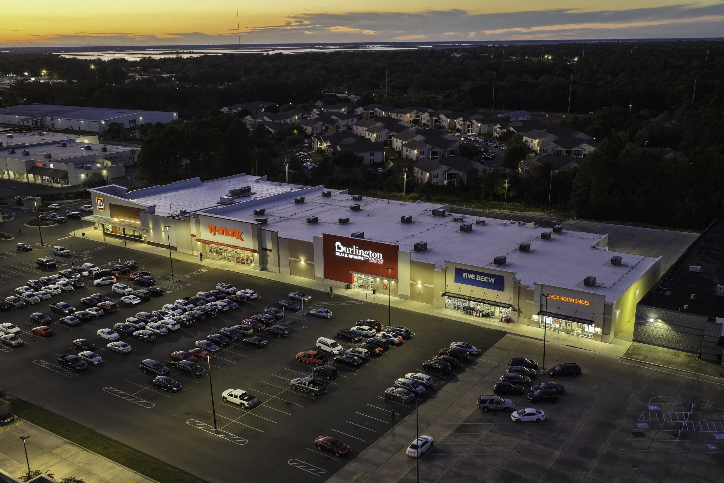 An aerial view of a shopping mall parking lot at dusk showing parked cars and illuminated stores, with residential homes and trees in the background.
