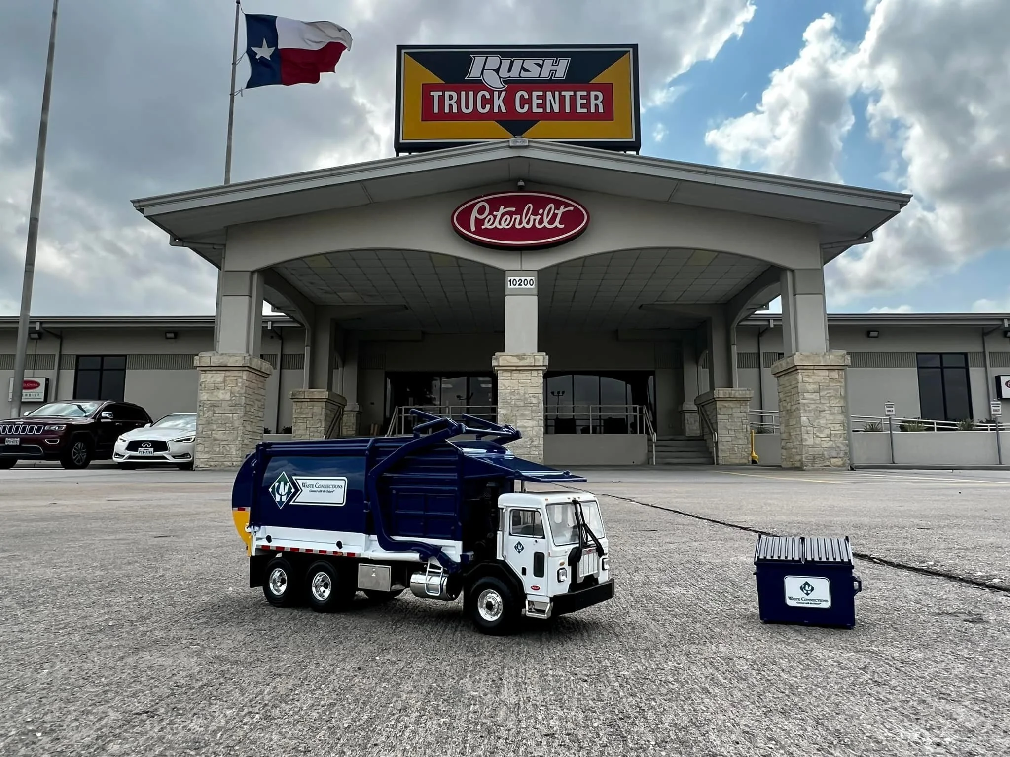A small toy garbage truck with a trash bin in front of a truck center building under a cloudy sky.
