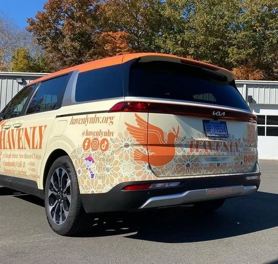 A minivan with religious and community organization branding, featuring an orange dove logo, website URL, and social media icons on a cream and orange background.