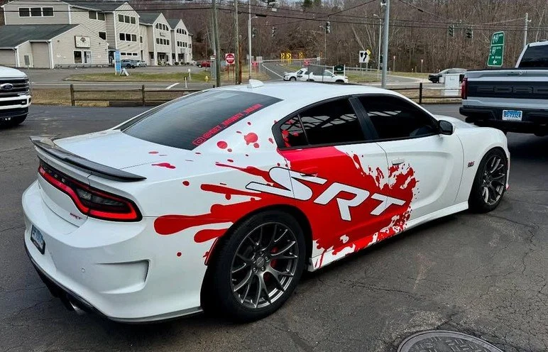 White sports car with red and black SRT decal and red splatter design on the side, parked in a lot with other vehicles and residential buildings in the background.