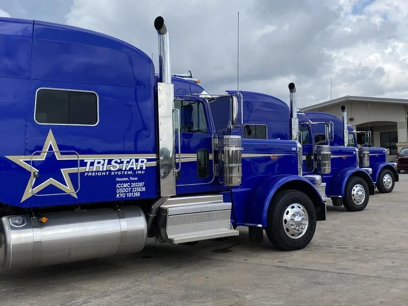 Three large blue semi-trucks parked in a row on a concrete lot, with a building and cloudy sky in the background.