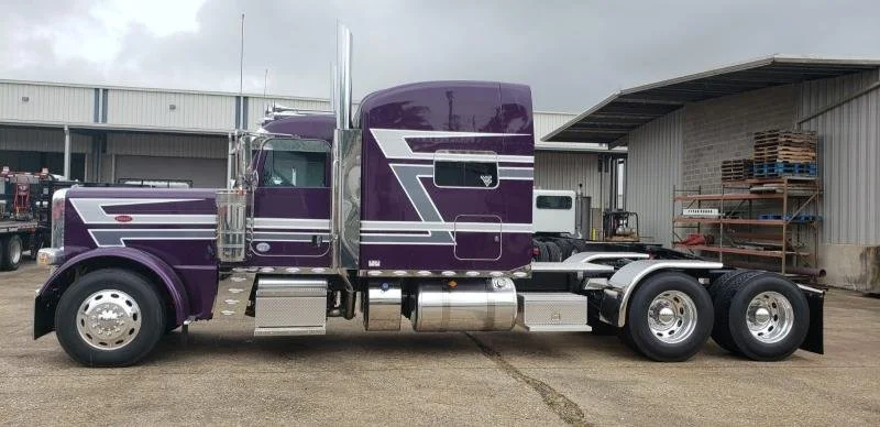 Purple semi-truck parked in front of a warehouse, with a large cab and six wheels.