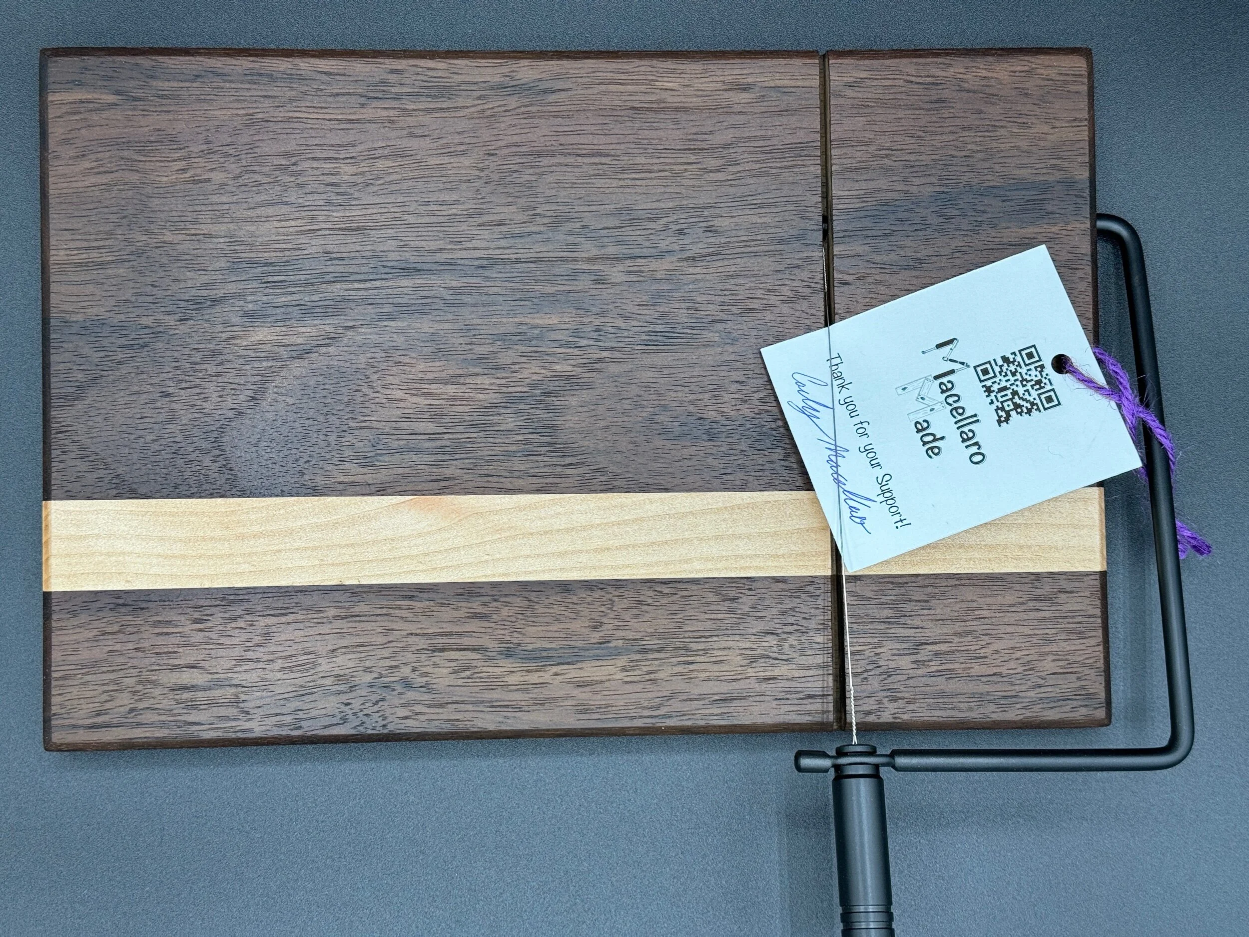 A wooden cheese slicer with a dark wood border and a strip of light wood in the center, placed on a gray surface. 