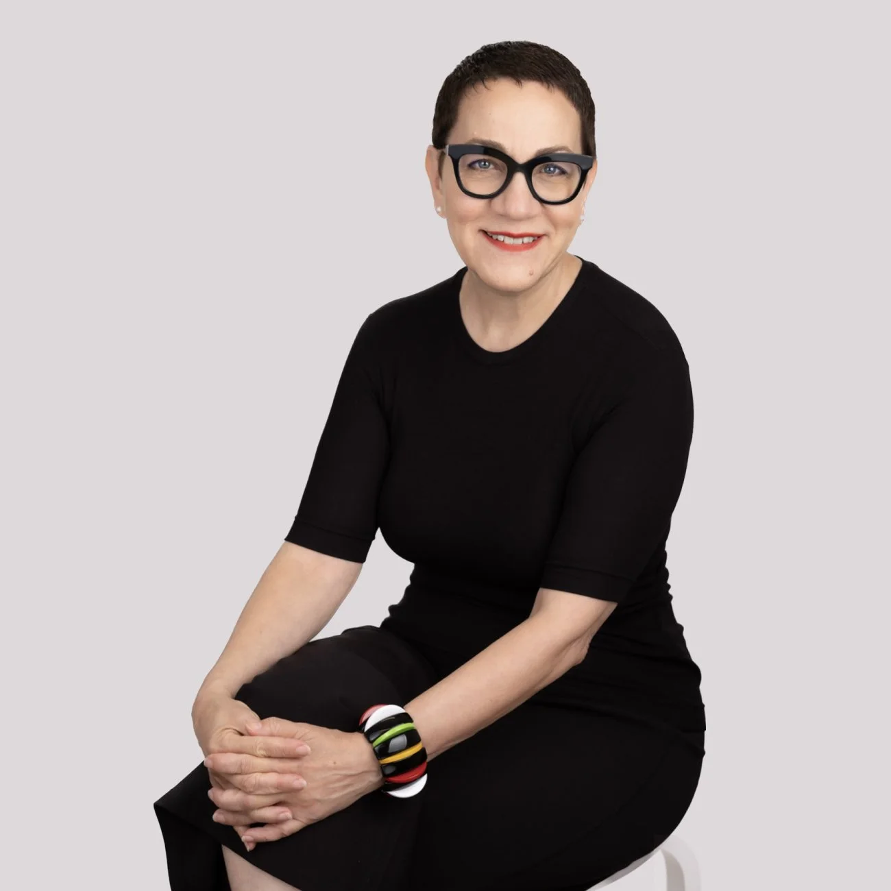 Portrait of Ann Webb, short dark hair, wearing glasses, a black top, and a colorful bracelet, sitting on a white stool against a plain gray background.