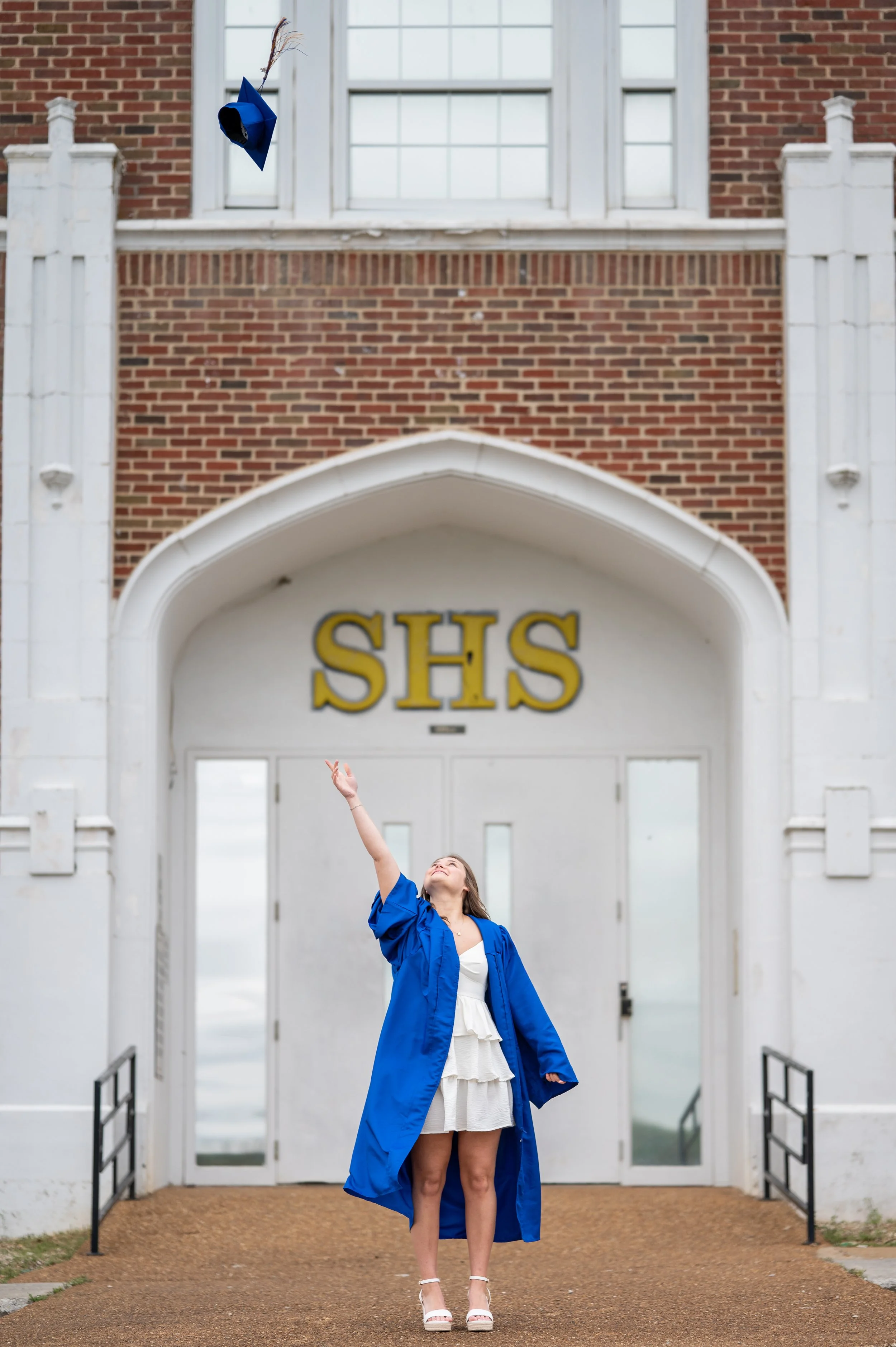 A young woman in a white dress and blue graduation gown standing in front of a school building, reaching up with her right hand to throw her cap into the air. The school building has red brick walls, white accents, and a sign with yellow letters read