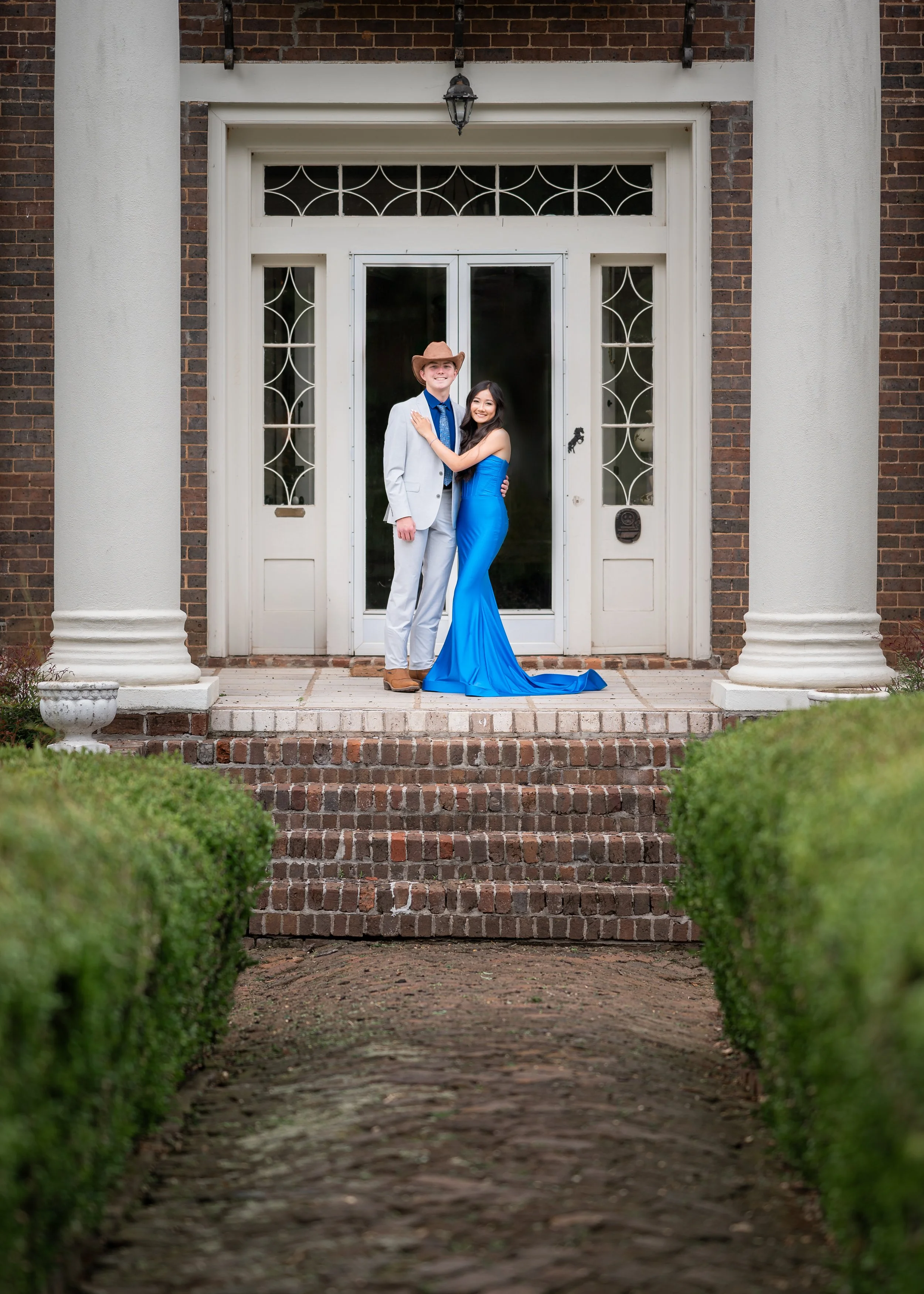A young couple dressed in formal attire standing on front porch steps of a brick house, smiling and embracing each other.