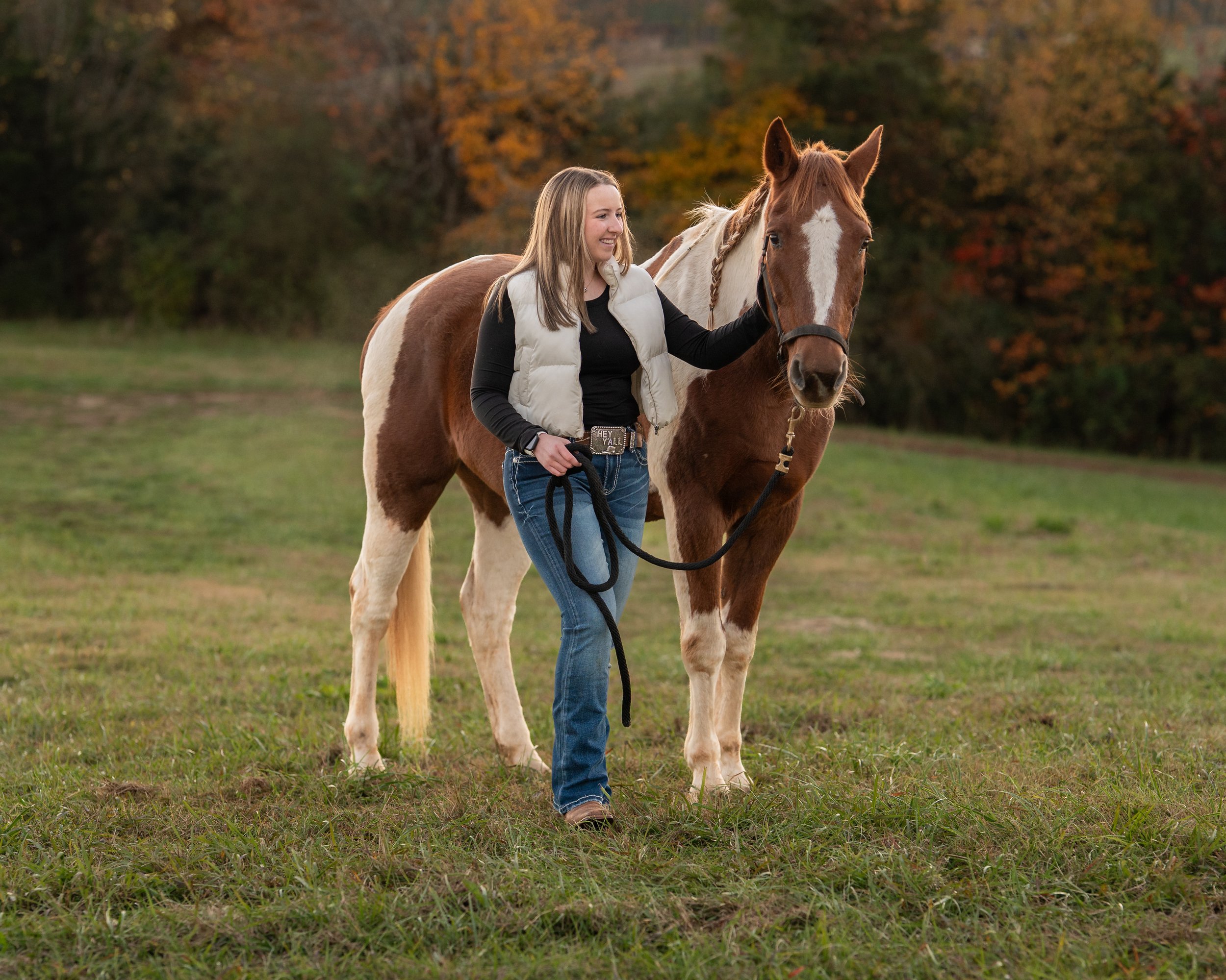 A woman walking a brown and white pinto horse in a grassy field with autumn trees in the background.