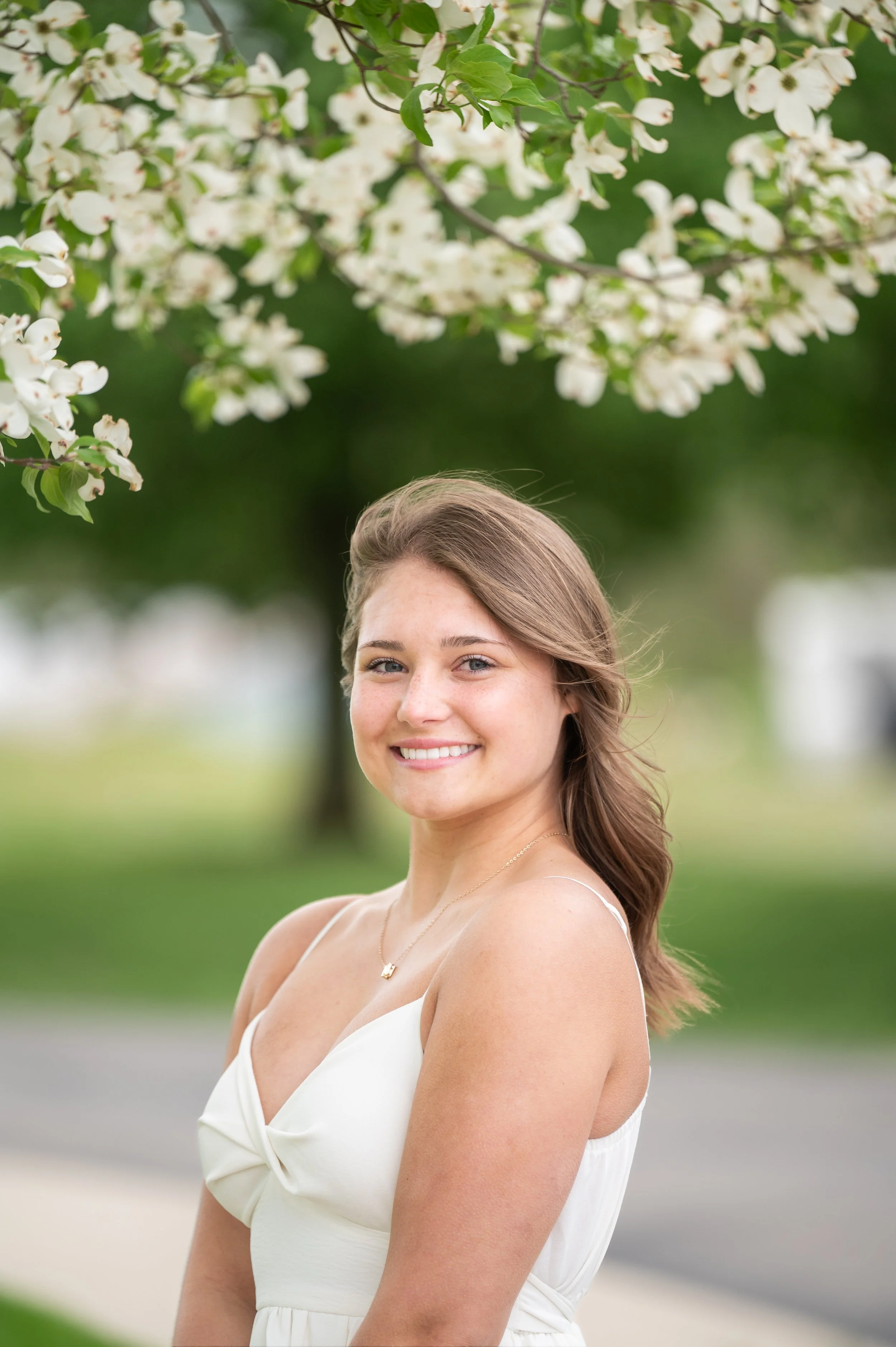 A young woman with long brown hair and a white dress standing outdoors under a blooming tree with white flowers, smiling at the camera.